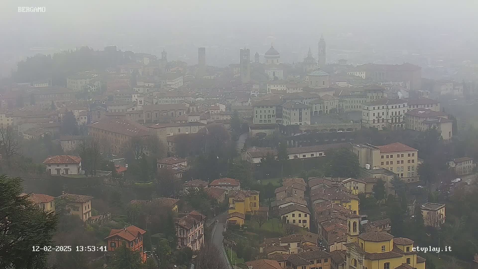 A foggy view of a city with numerous buildings, including churches and tall structures, stretches out from a high vantage point.