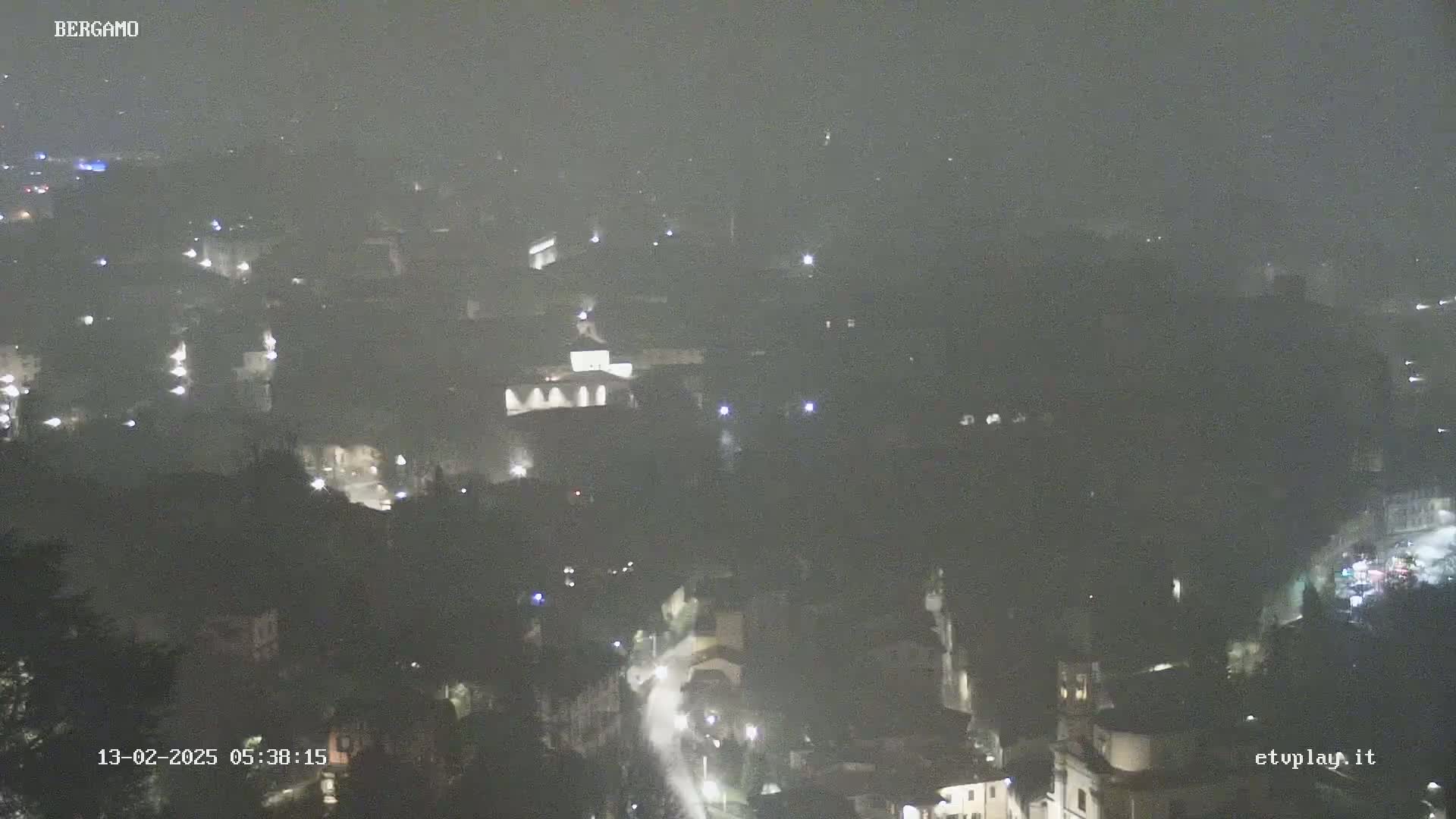 A nighttime, hazy view of a city, possibly under light rain or mist, shows scattered lights and buildings.