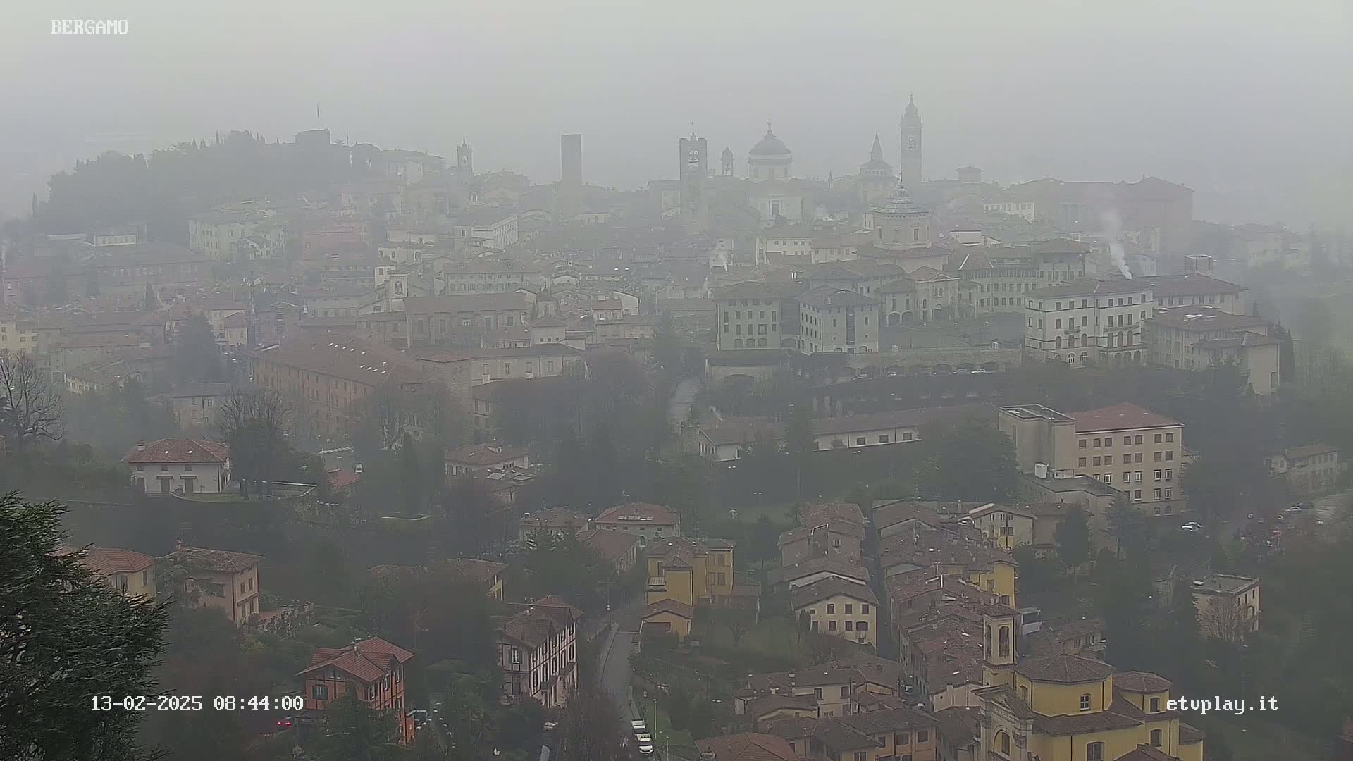 A sprawling city, shrouded in a thick fog, is viewed from a high vantage point, showing numerous buildings and architectural features.