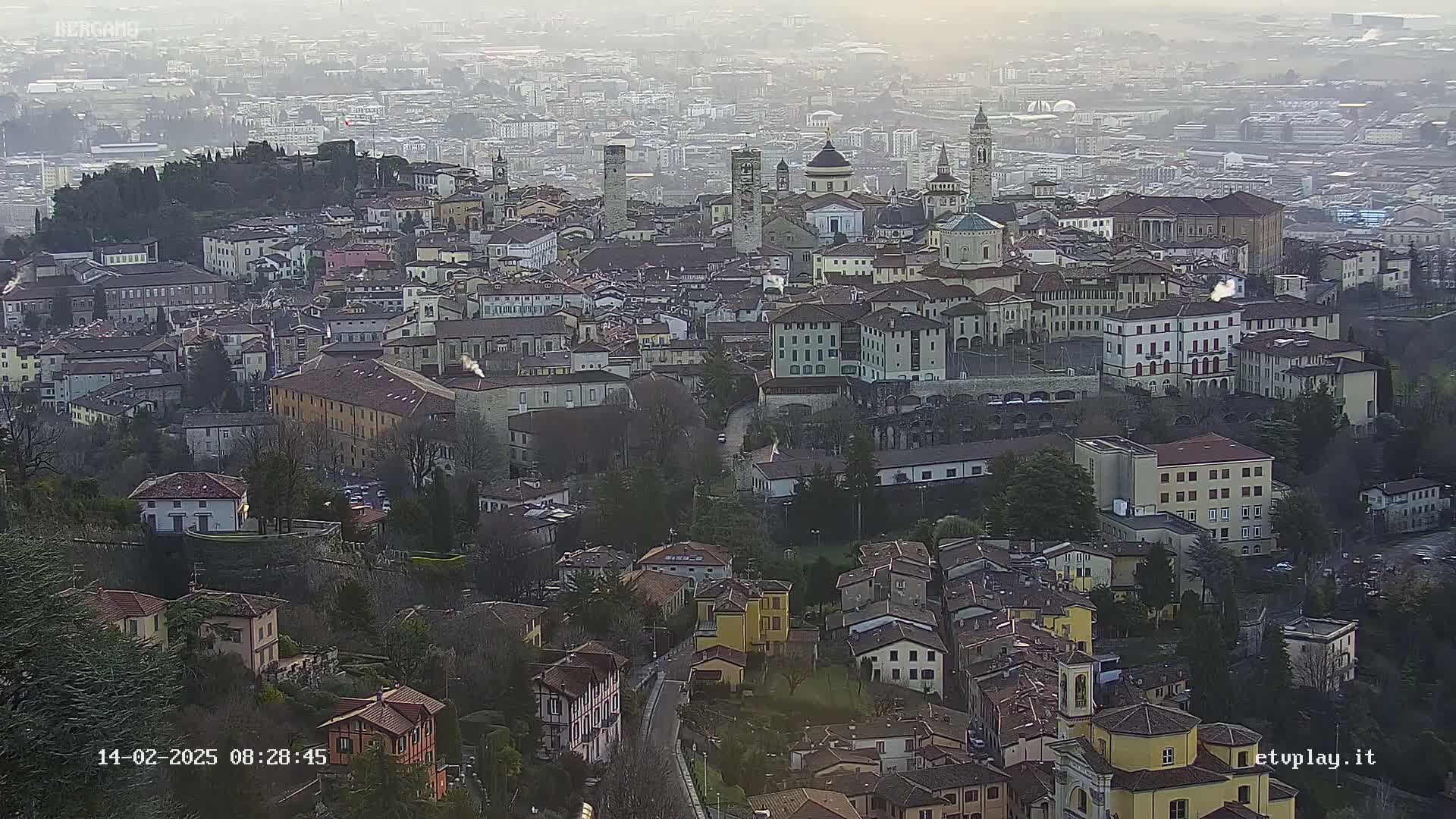 A hazy, distant view of a sprawling city with numerous buildings of varying heights and architectural styles, nestled among hillsides and greenery.