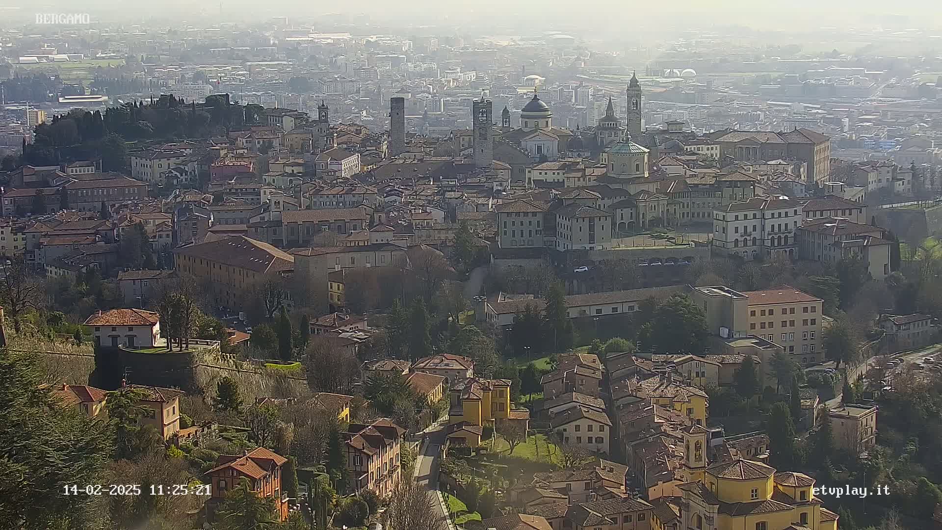 A hazy, sunny day view of a sprawling city with many historic buildings, including a large cathedral and several towers, nestled among hills.