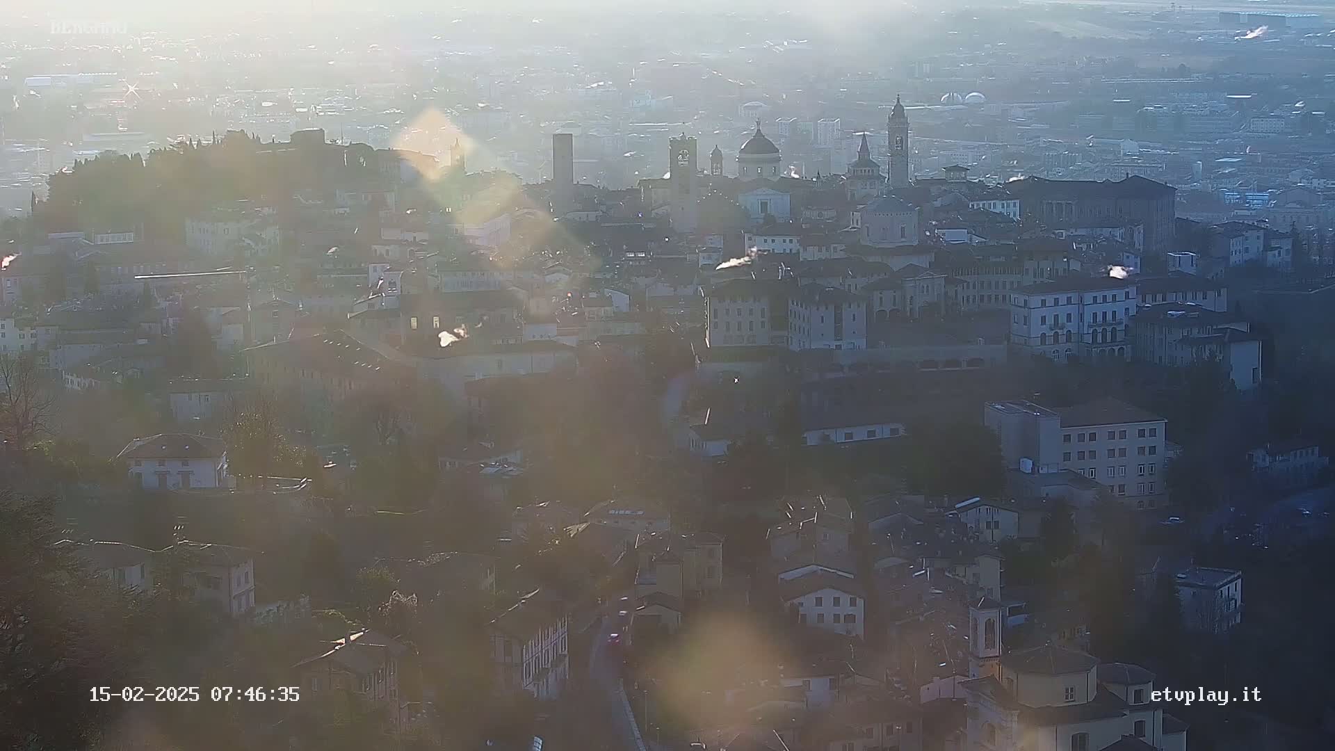 A hazy, sunlit view of a European city sprawling across a hillside, with numerous buildings and churches visible.