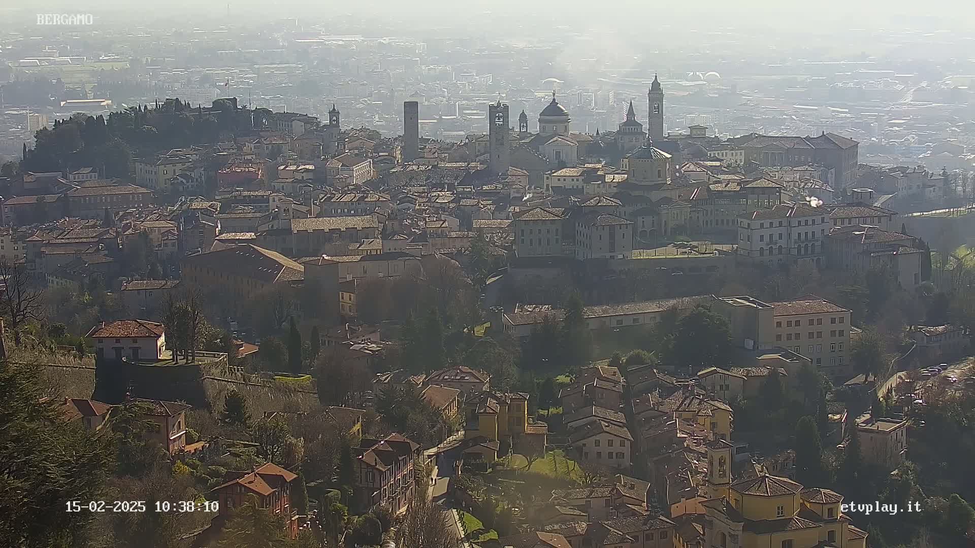 A hazy, sunny day overlooks a sprawling cityscape of terracotta-roofed buildings, punctuated by several prominent towers and churches.