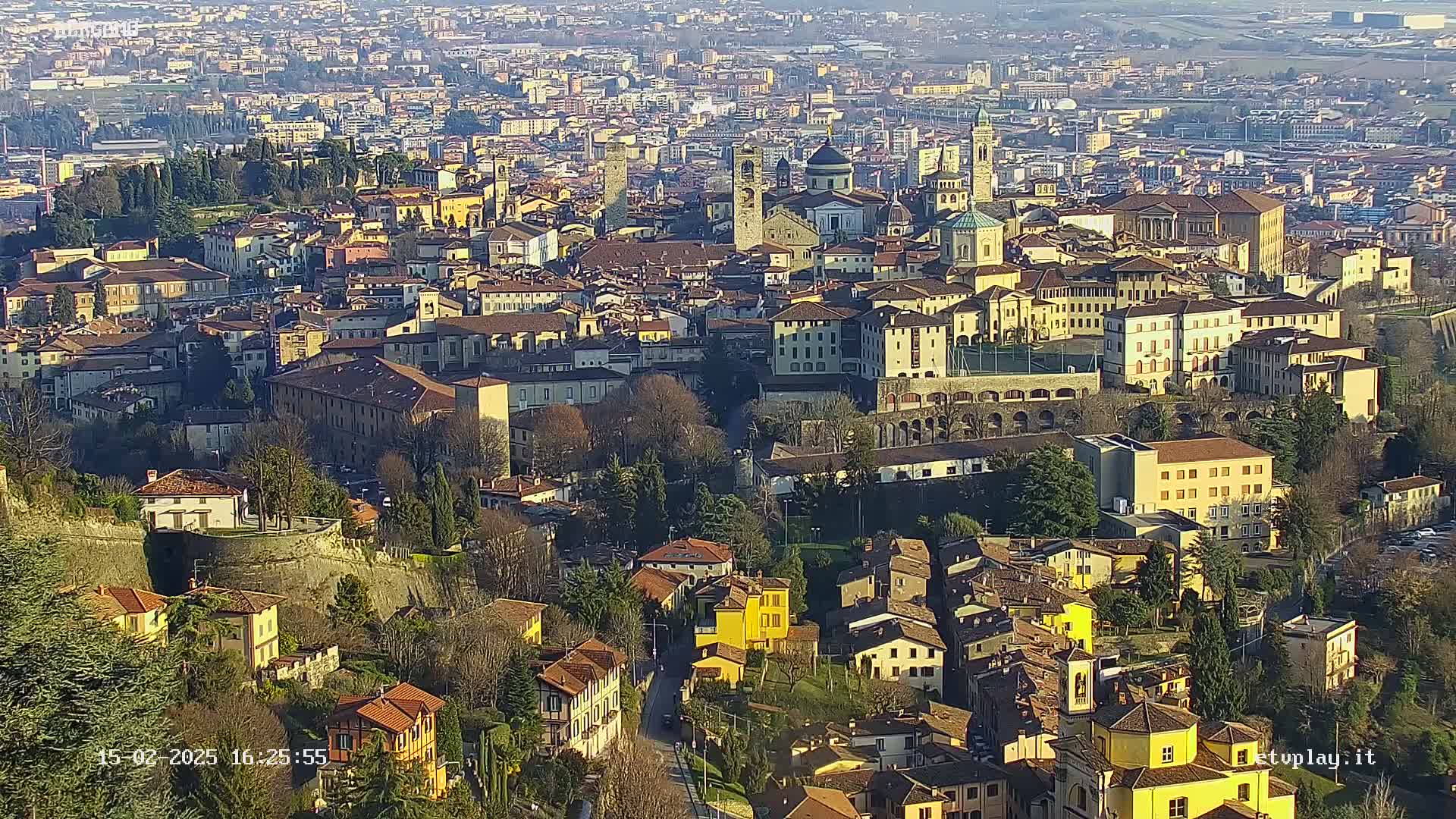 An aerial view of a densely packed European city on a sunny day, showing many buildings of varying heights and architectural styles, surrounded by trees and hills.