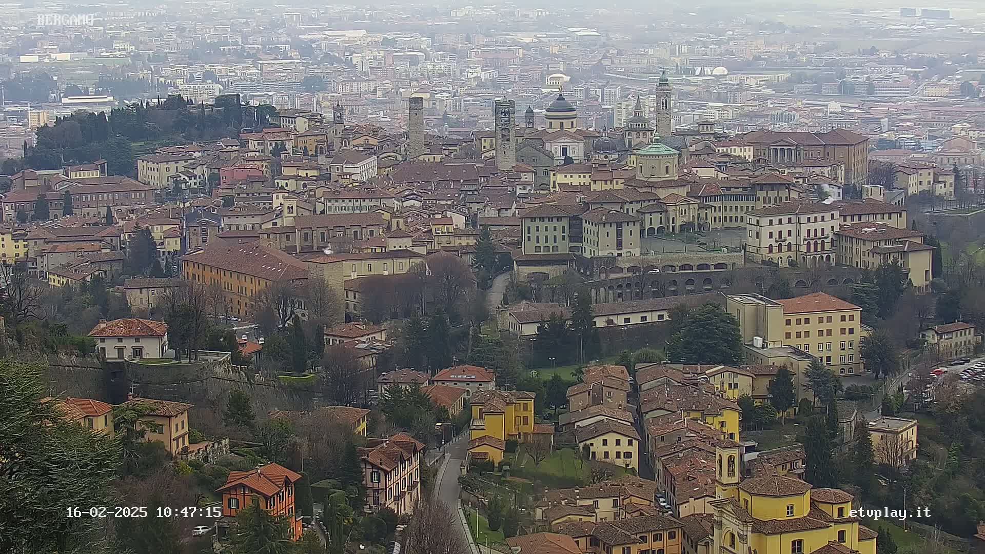 A sprawling city with numerous terracotta-roofed buildings, churches, and a surrounding landscape of trees under an overcast sky.