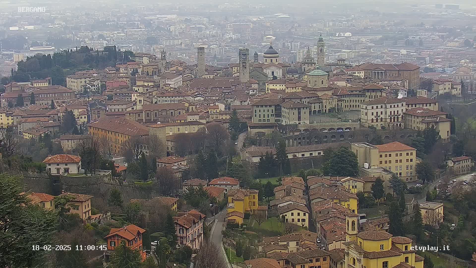 A sprawling city with numerous brown-roofed buildings, some with towers and domes, is visible under an overcast sky.