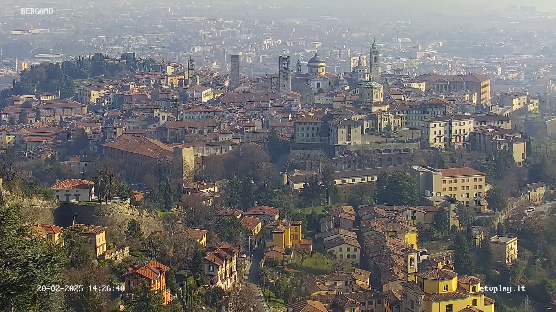 A hazy, sunlit view of a sprawling city with numerous terracotta-roofed buildings, including several prominent churches and towers.