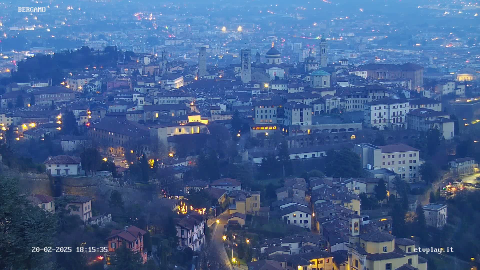 A sprawling city at twilight, illuminated by scattered streetlights, is nestled in a valley under a hazy sky.