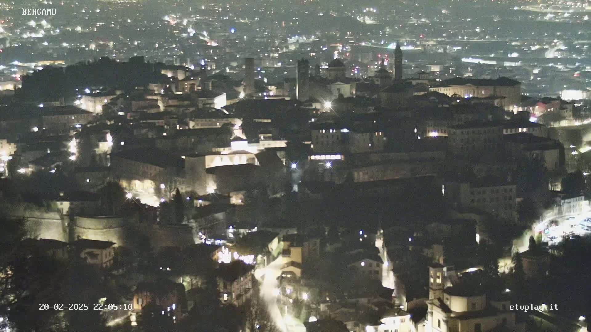 A nighttime aerial view of a city with many buildings illuminated, nestled in a hilly landscape.