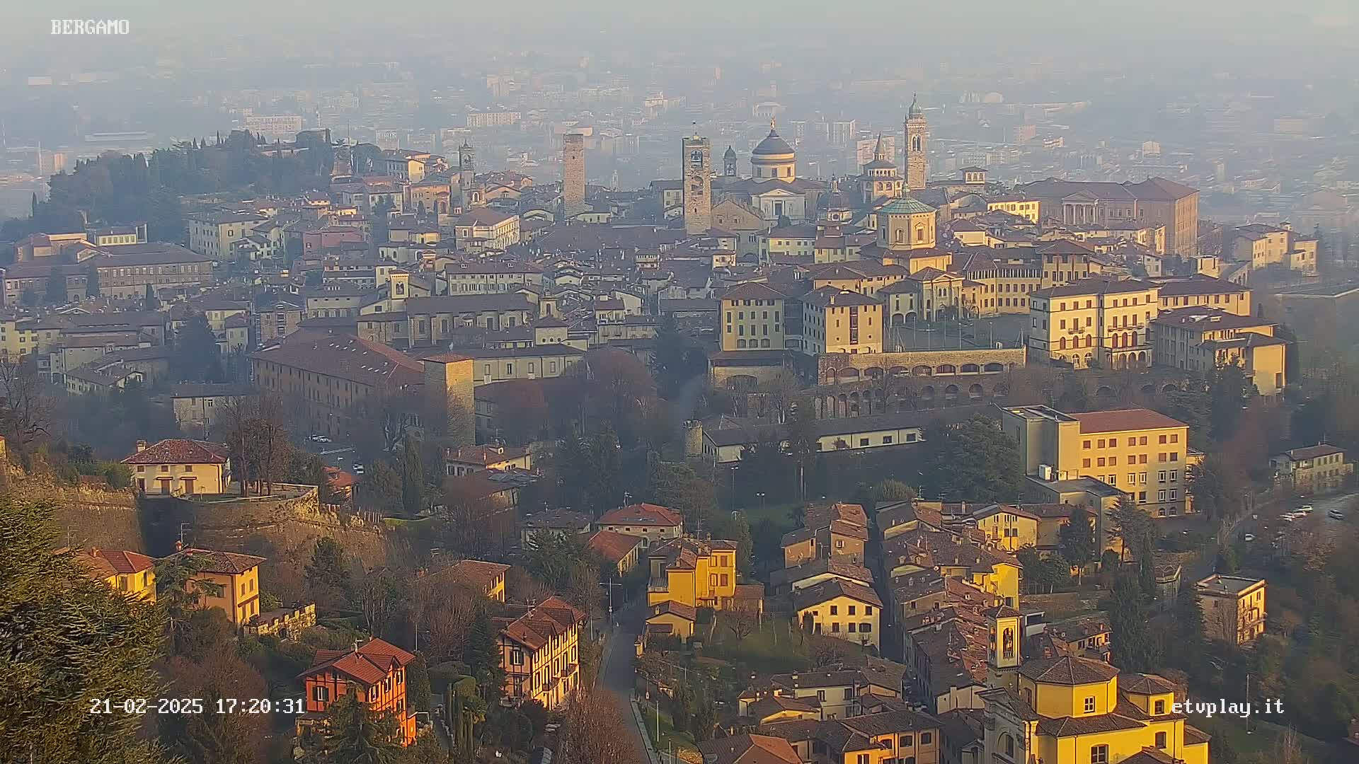 A hazy, sunlit view of Bergamo, Italy, showing numerous buildings densely clustered together on a hillside.