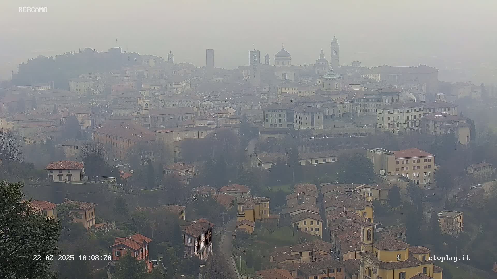 A hazy view of a sprawling city with numerous buildings, including several churches and towers, is seen from an elevated vantage point.