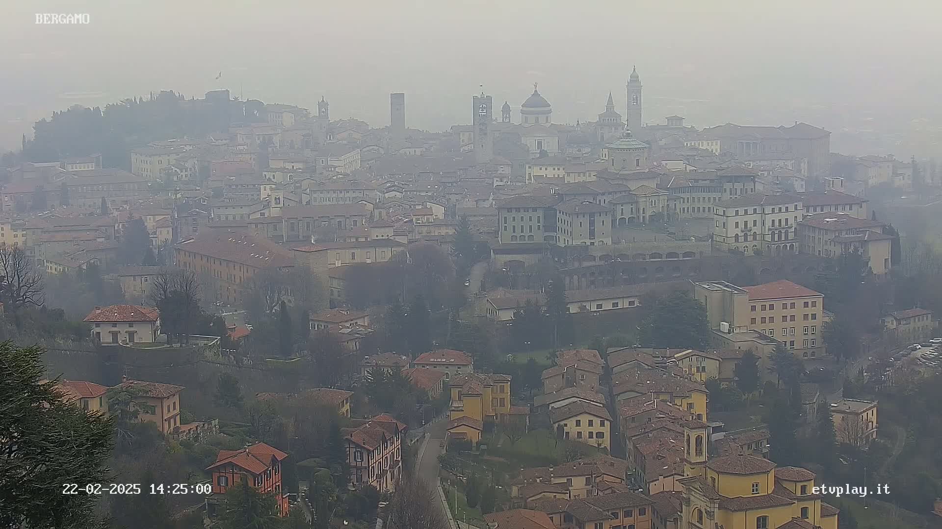 A hazy view of a city with numerous buildings, including churches and towers, set against a backdrop of hills.