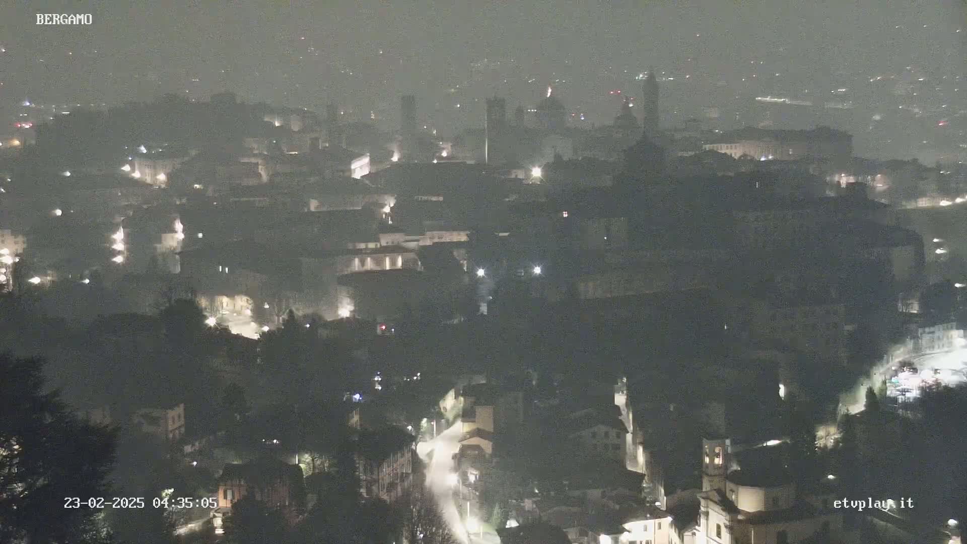 A nighttime, hazy view of a city with dimly lit buildings and roads winding through hills.