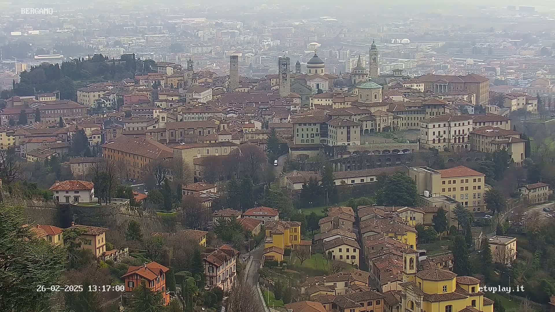 A hazy view of a sprawling city with numerous brown-roofed buildings, including churches and taller structures, nestled within a landscape of trees and greenery.