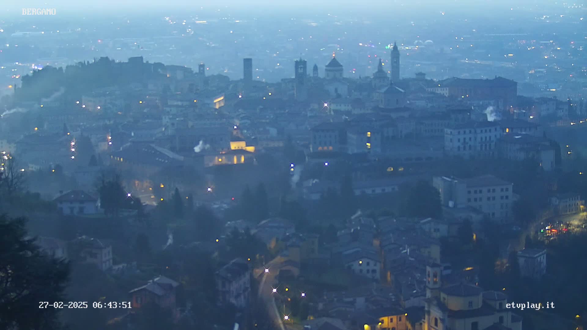 A hazy, dimly lit cityscape at dawn, with numerous buildings and scattered lights.