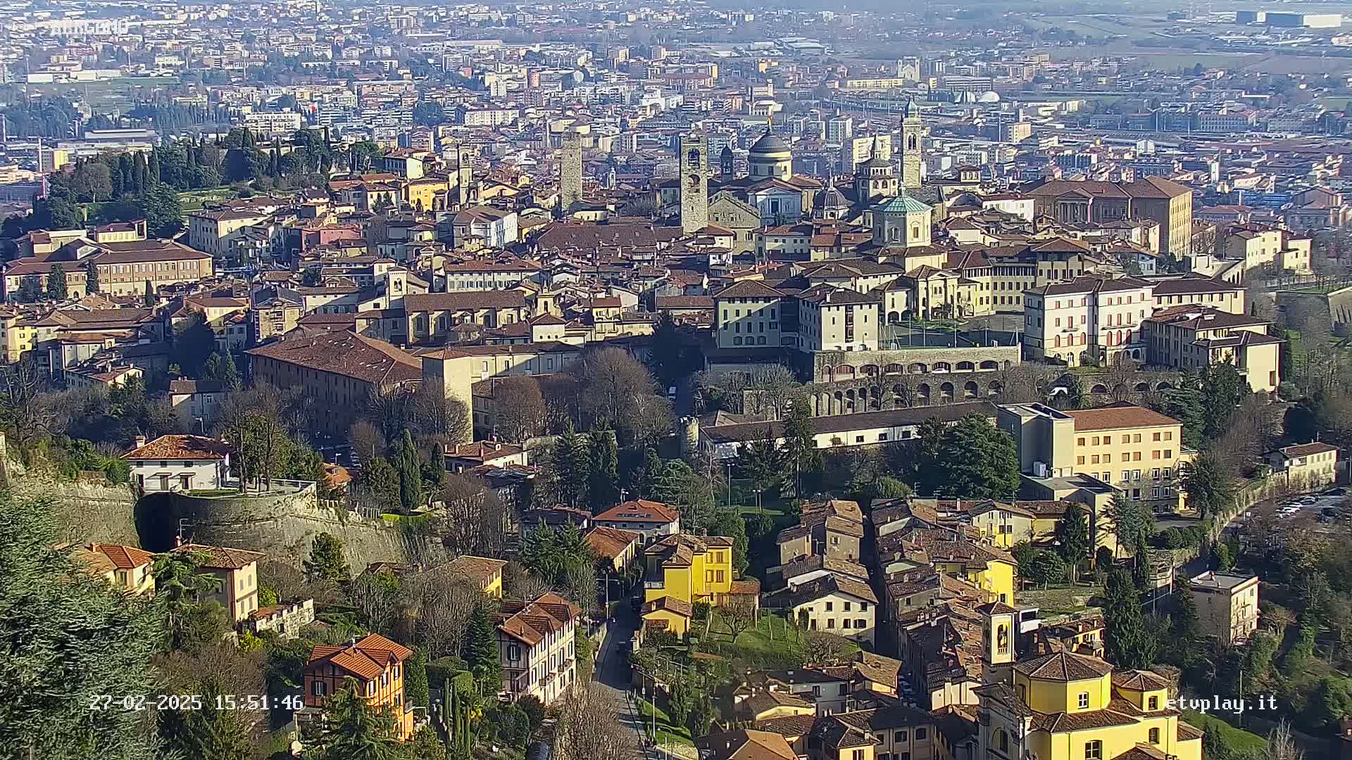 A sprawling city with many tan and brown buildings, some with terracotta roofs, is visible from a high vantage point on a sunny day.