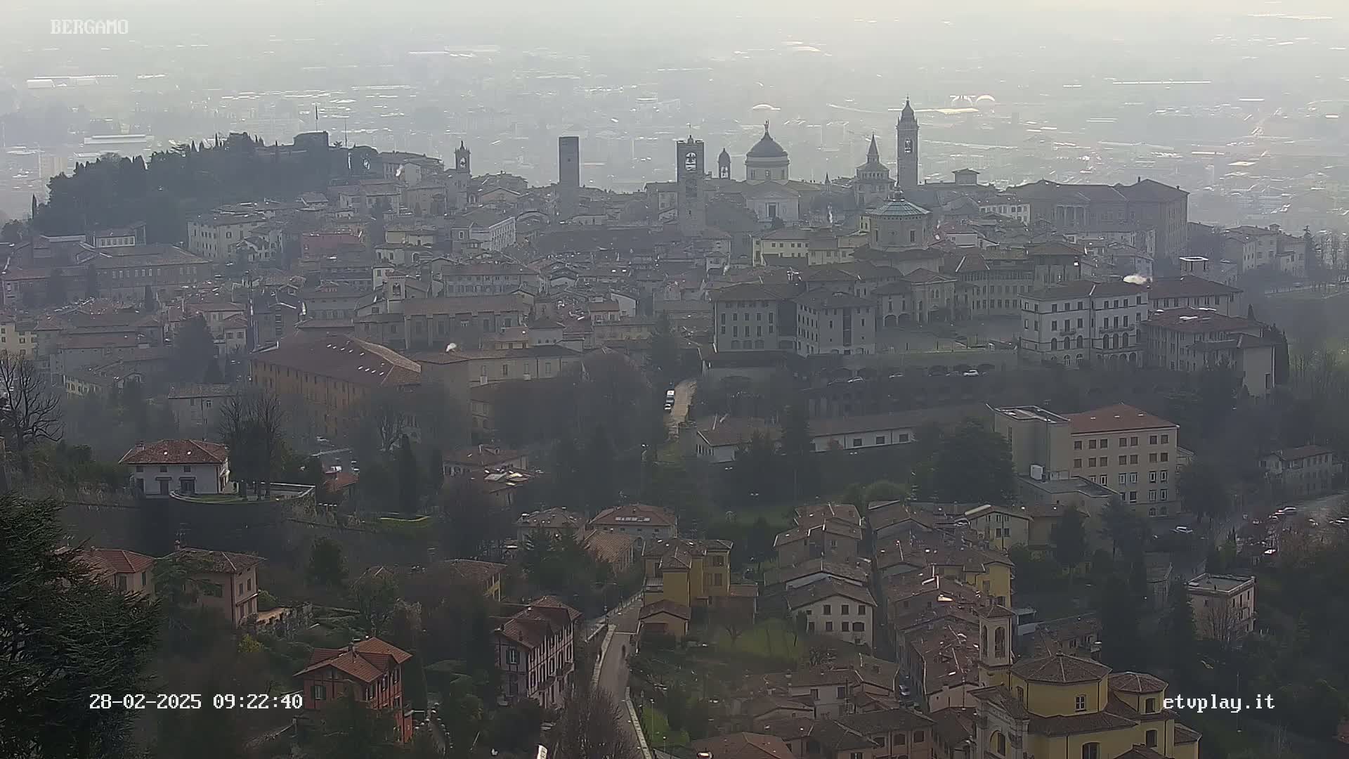 A hazy, aerial view of a sprawling city with numerous buildings and several prominent towers and domes, suggesting a historical European city.
