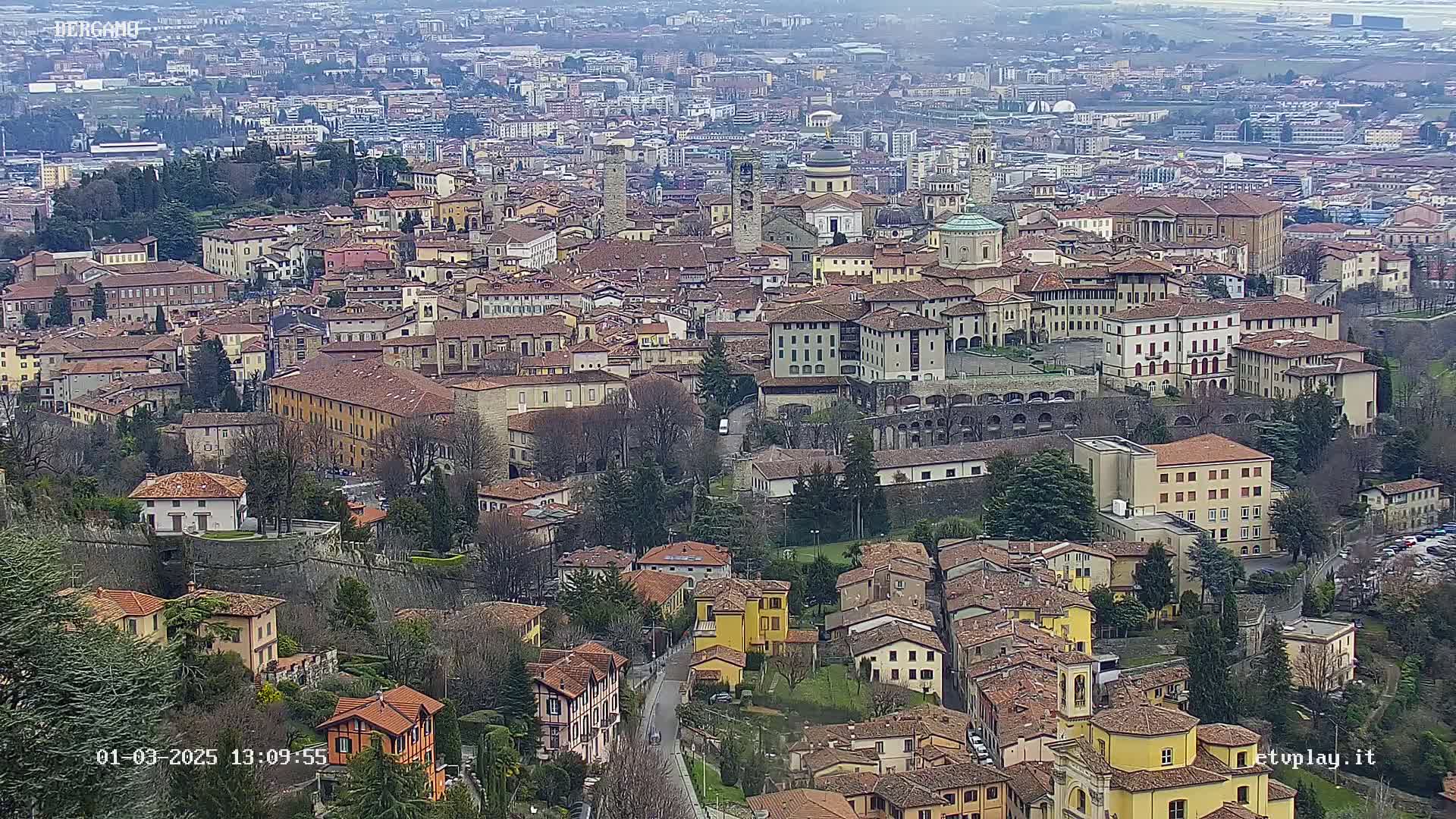 An expansive view of a city with numerous brown-roofed buildings, nestled within a valley on an overcast day.
