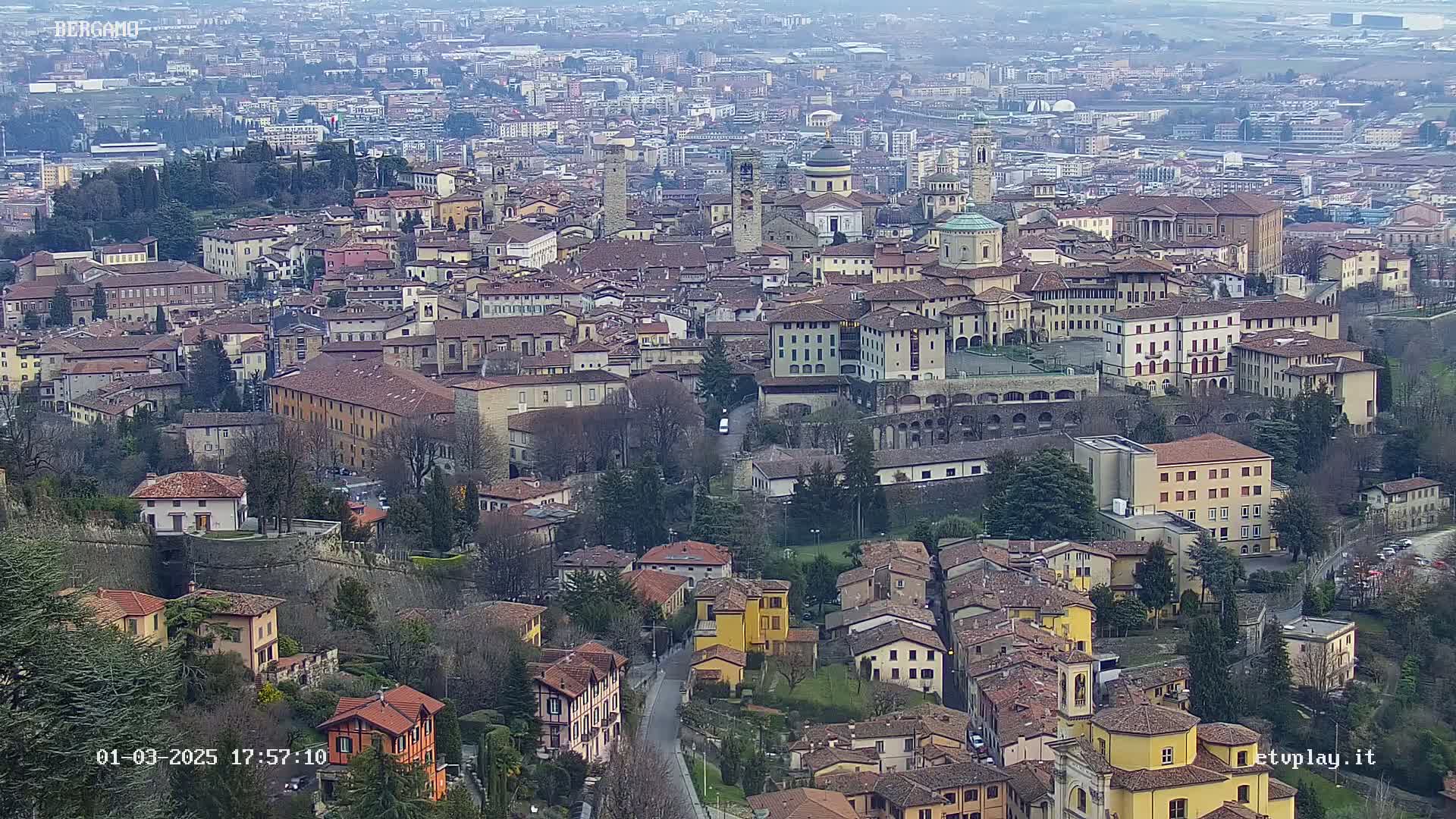 A sprawling city with numerous buildings of varying heights and architectural styles, nestled within a landscape of greenery under an overcast sky.