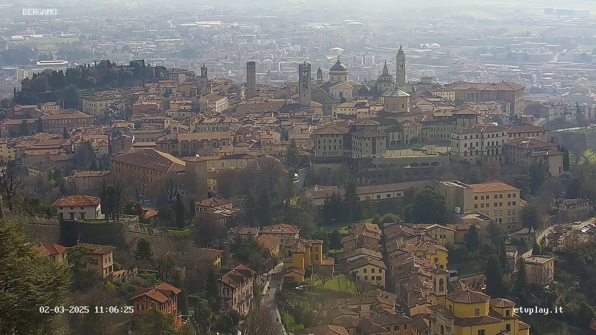 A hazy, sunny day reveals a sprawling cityscape of terracotta-roofed buildings, interspersed with churches and towers, nestled in a valley.