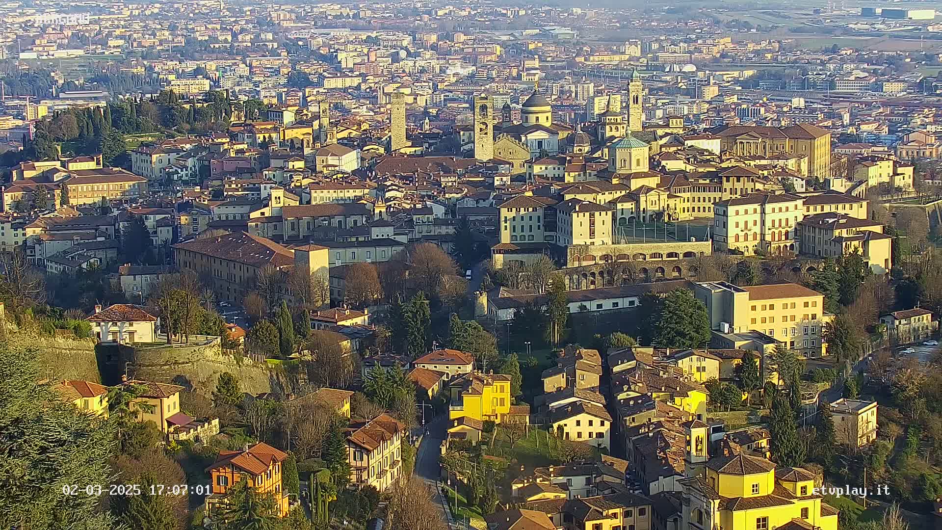 An aerial view of a city with many buildings of similar style and color, situated on a hillside, under a mostly sunny sky.