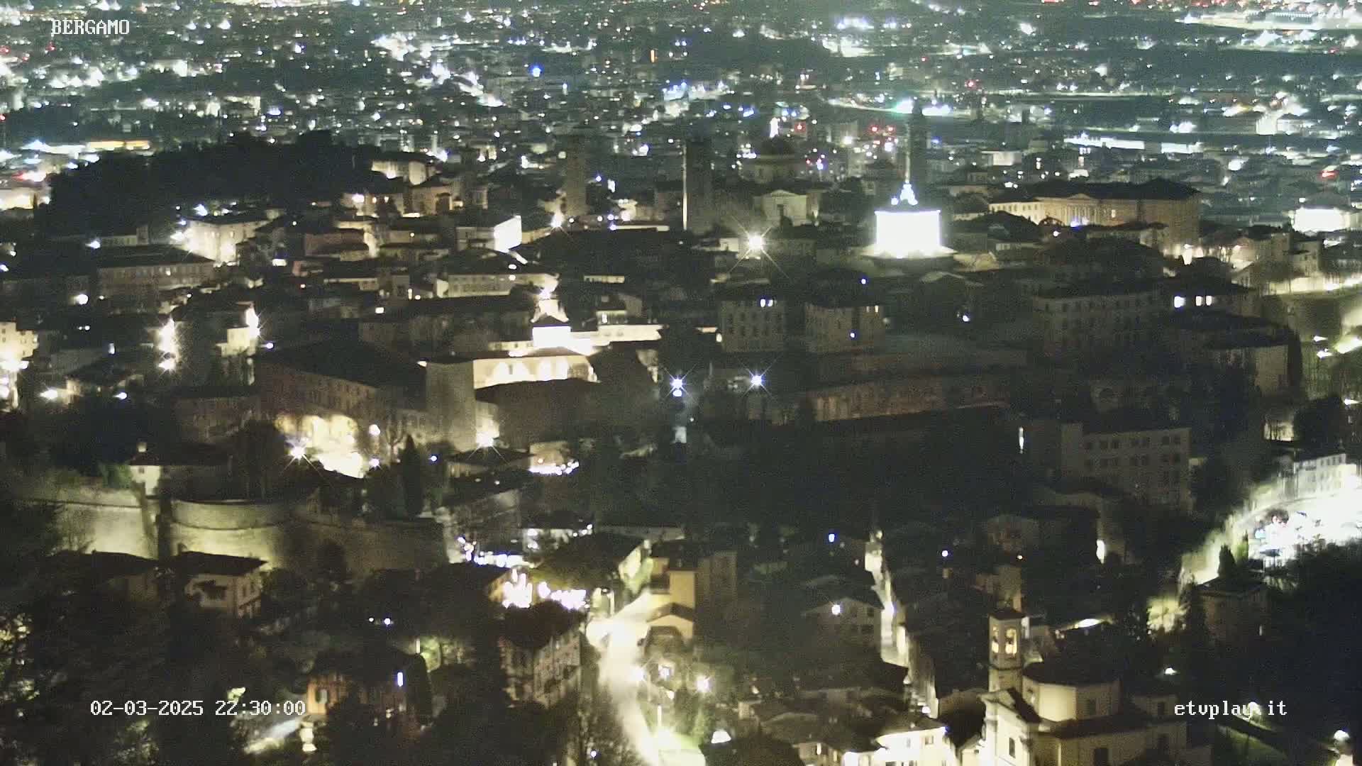A nighttime aerial view of a city illuminated by streetlights, showing a dense collection of buildings nestled within walls.