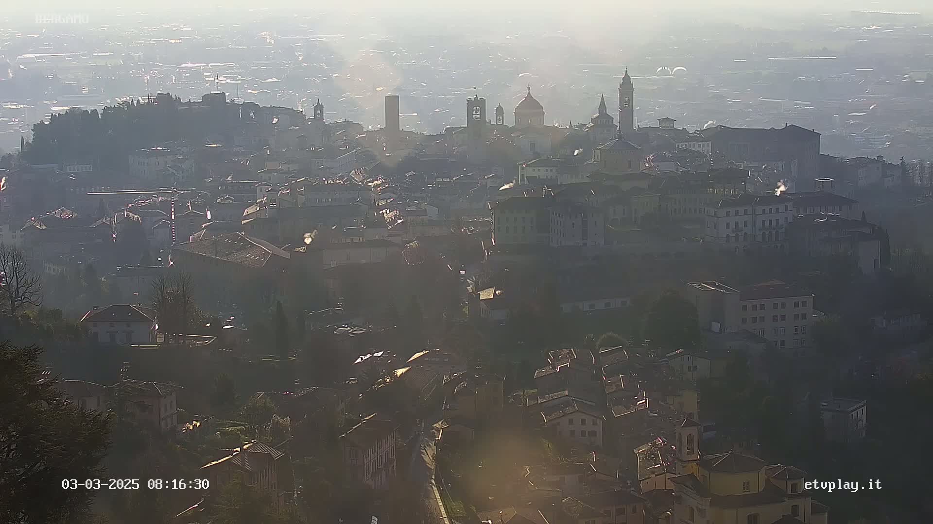 A hazy, sunlit view of a city sprawling across a hillside, with numerous buildings and church spires visible in the distance.