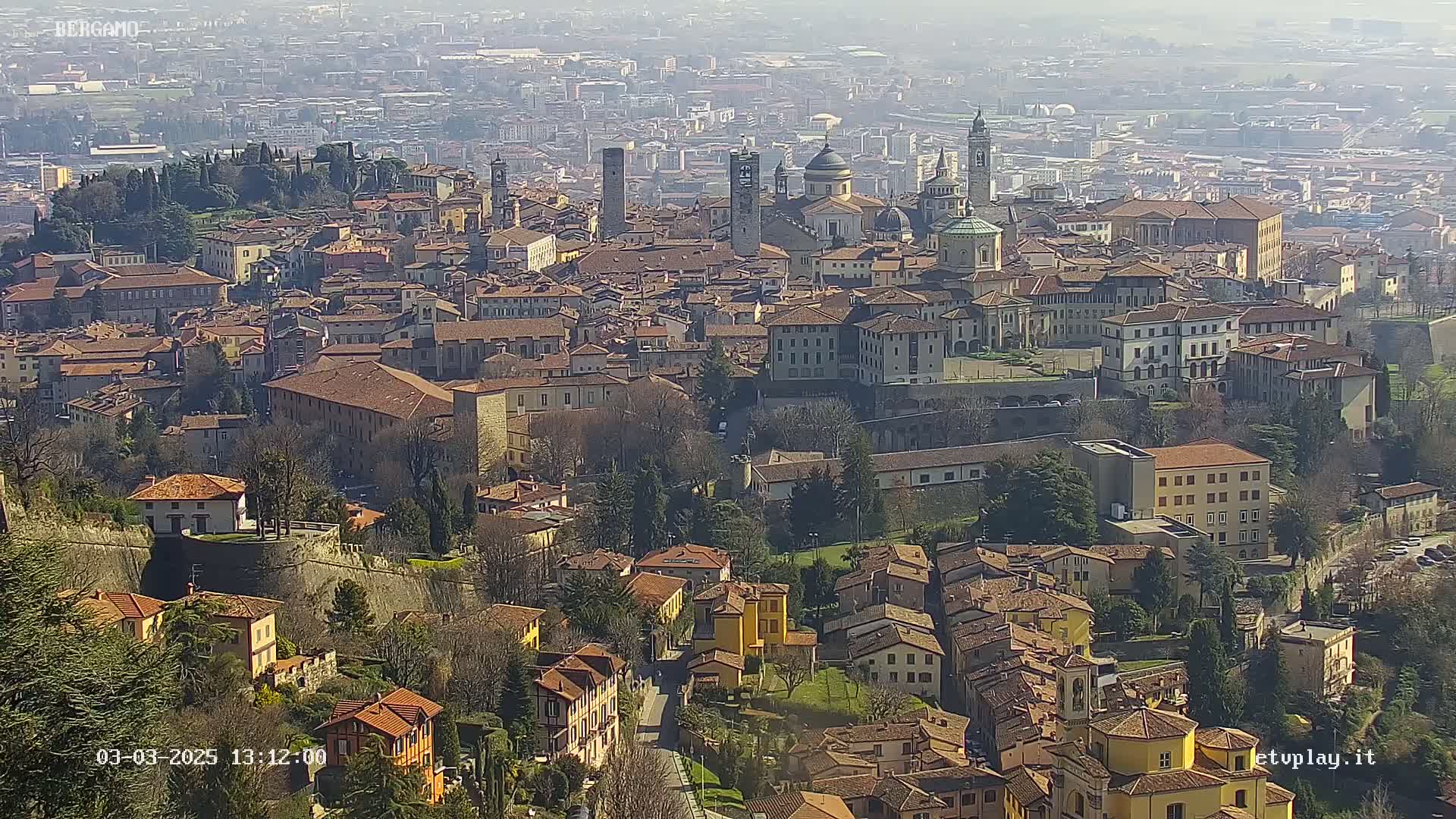 A sprawling city of terracotta-roofed buildings nestled in a valley on a sunny day, with hazy mountains visible in the distance.
