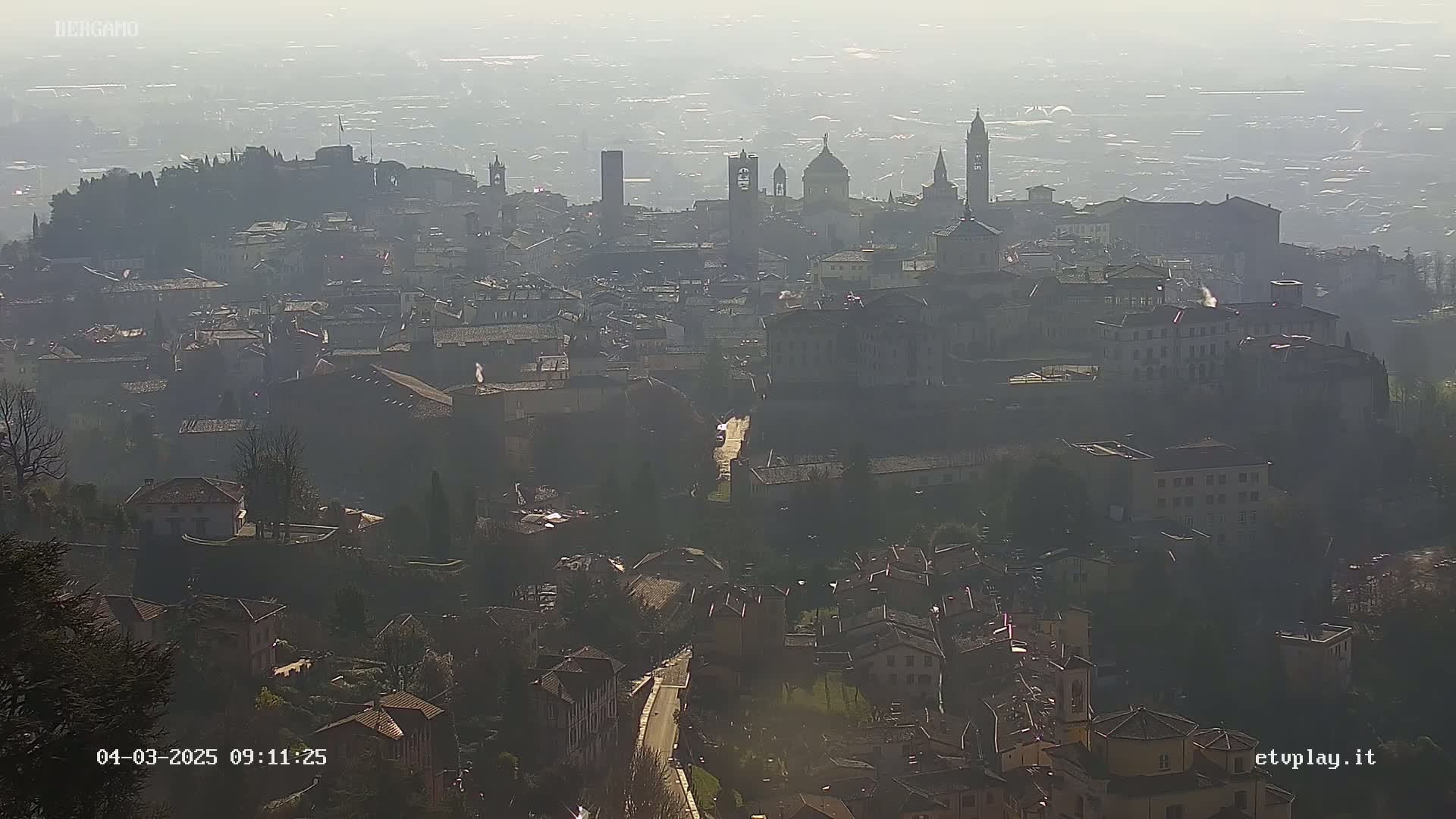 A hazy view of a city with numerous buildings and towers, some appearing to be churches, on a seemingly sunny day.