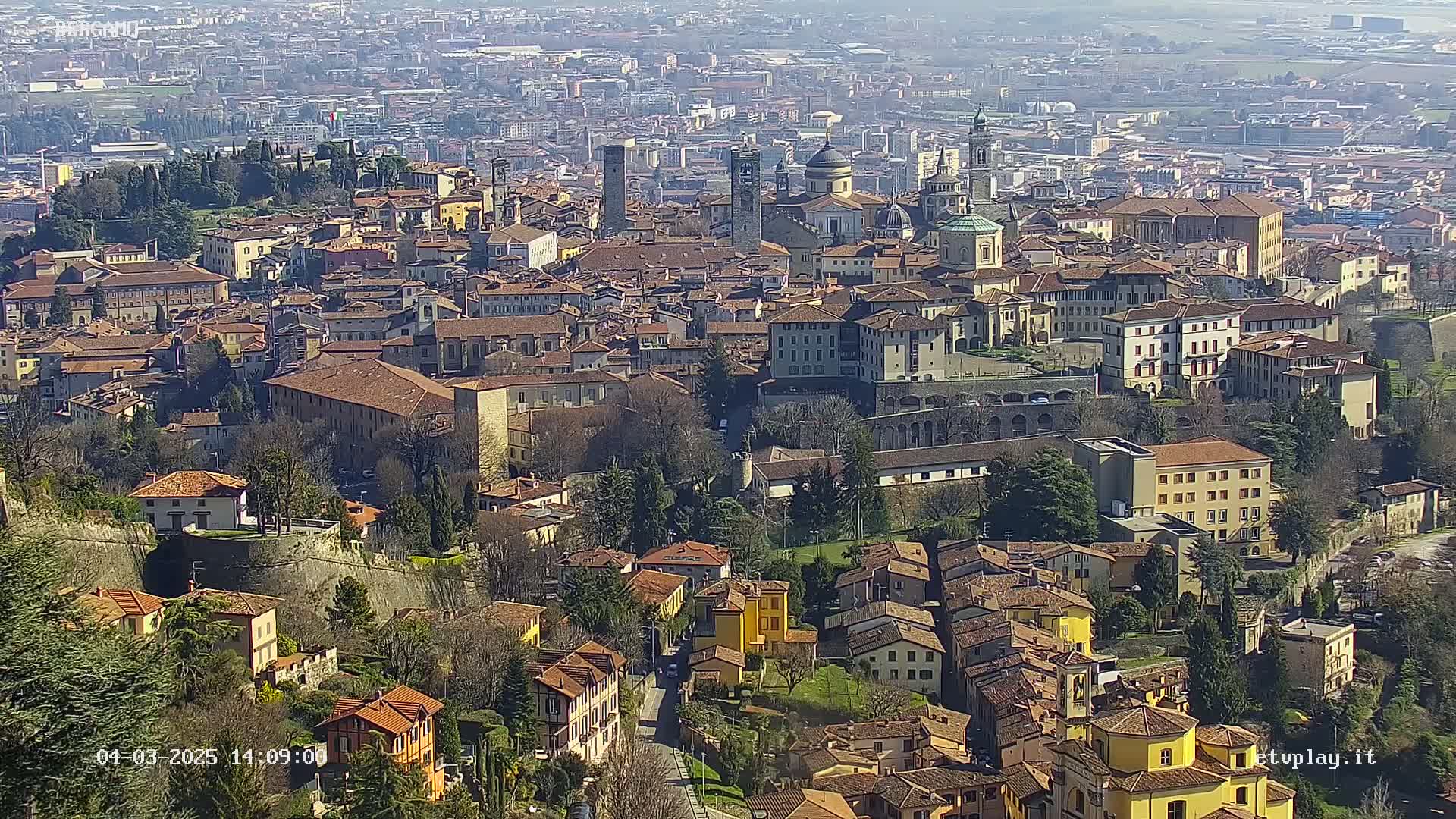 A sprawling city with many brown-roofed buildings, surrounded by walls and green spaces, under a clear sunny sky.