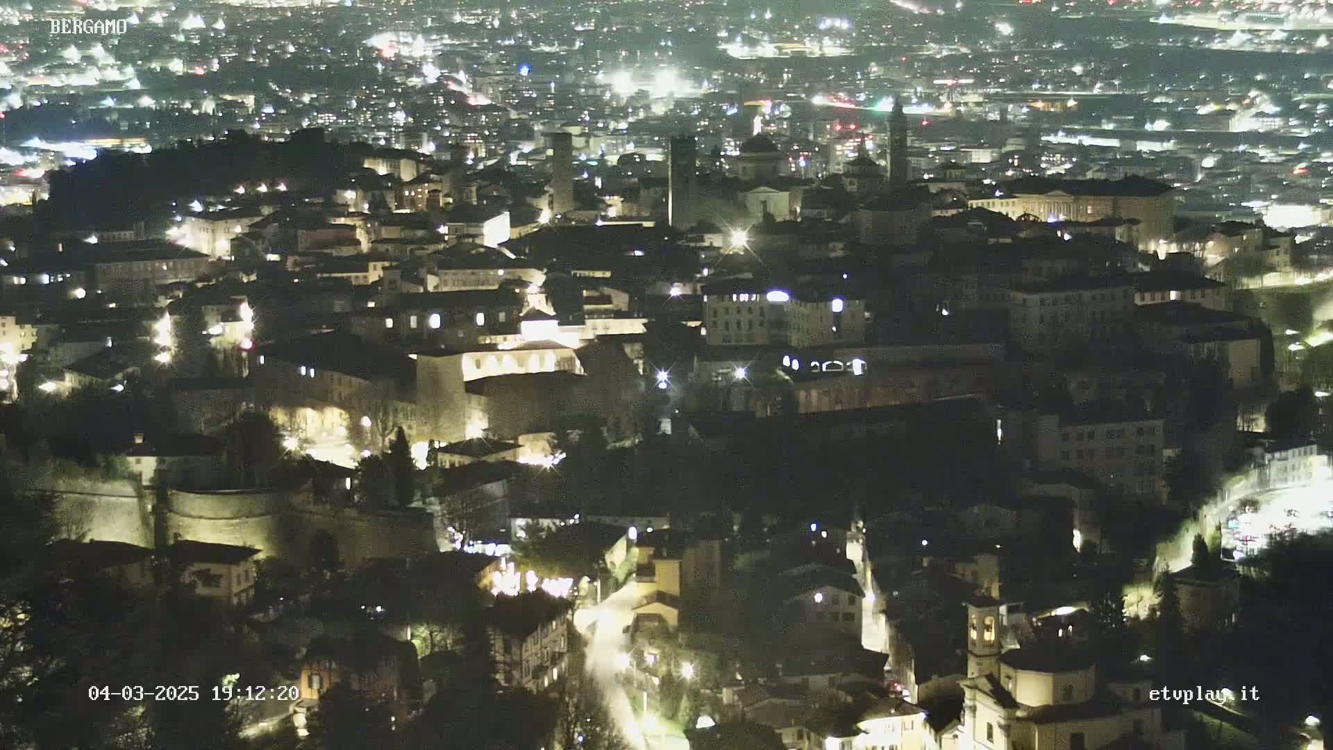 A nighttime aerial view of a city, showing many illuminated buildings nestled together within the confines of ancient-looking walls.