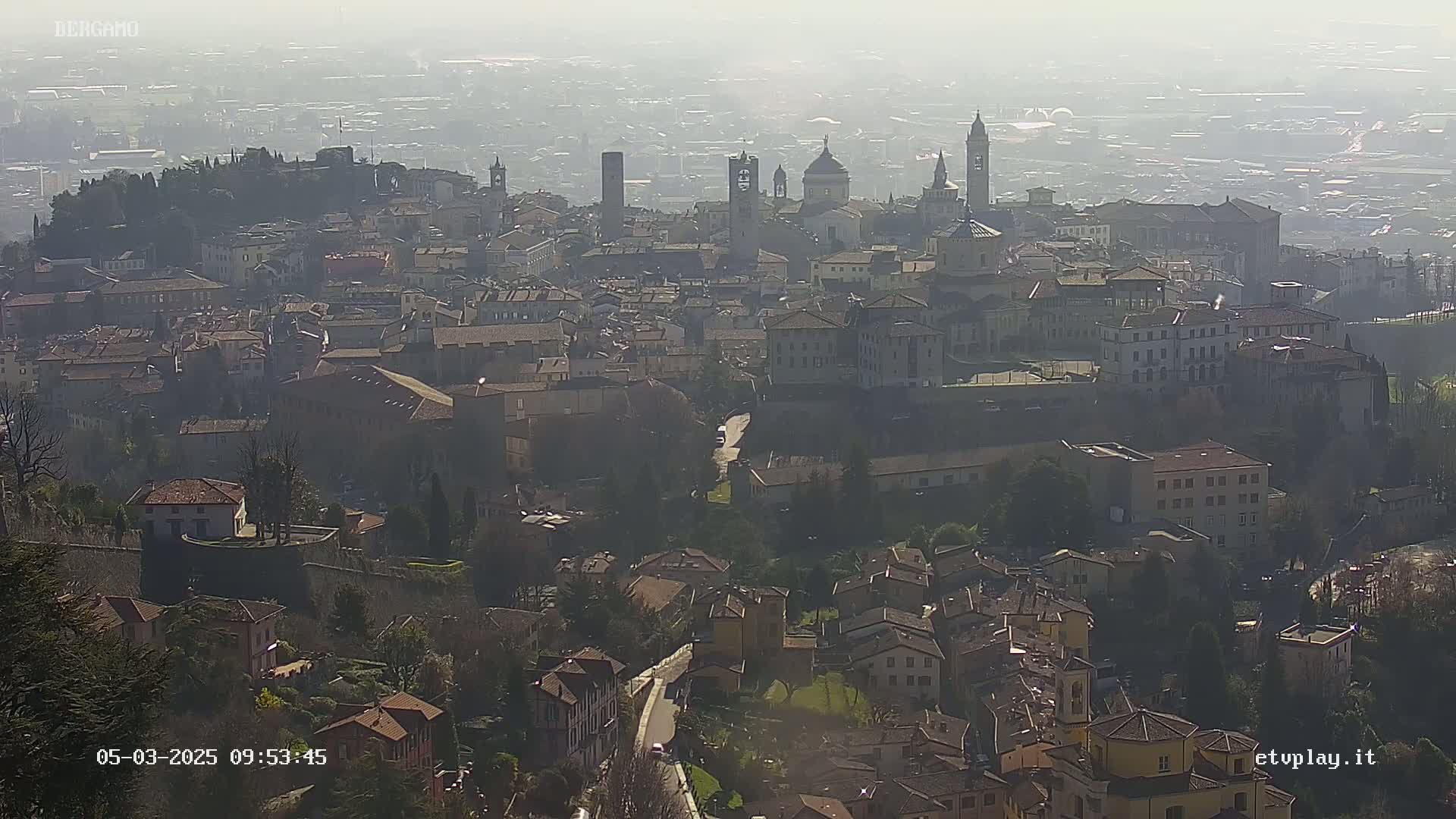 A hazy, sunny view of a sprawling city with numerous buildings, including churches and towers, nestled among hills.