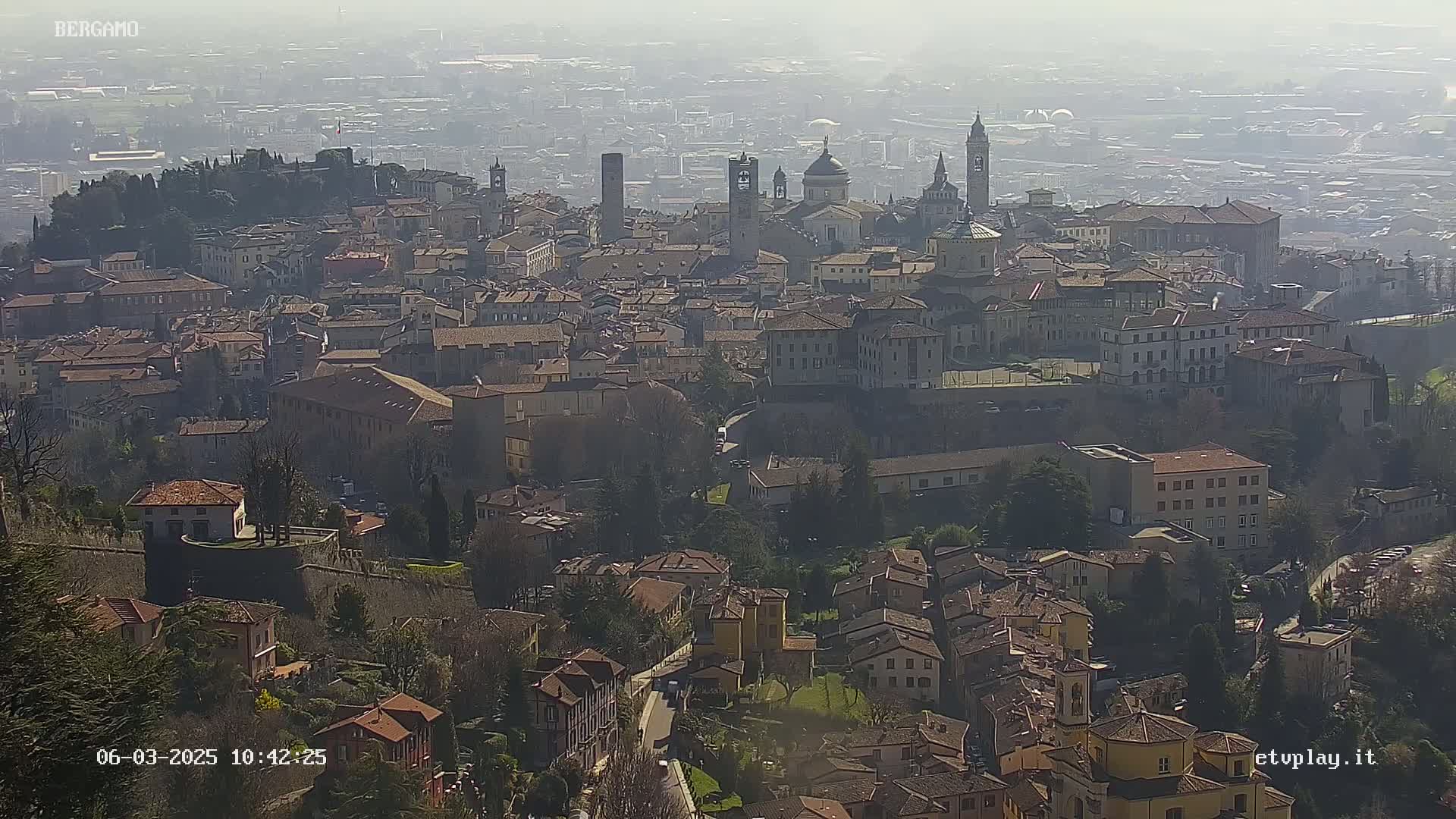 A hazy, sunlit cityscape of many closely packed buildings, including churches and a fortress, stretches across a hill.