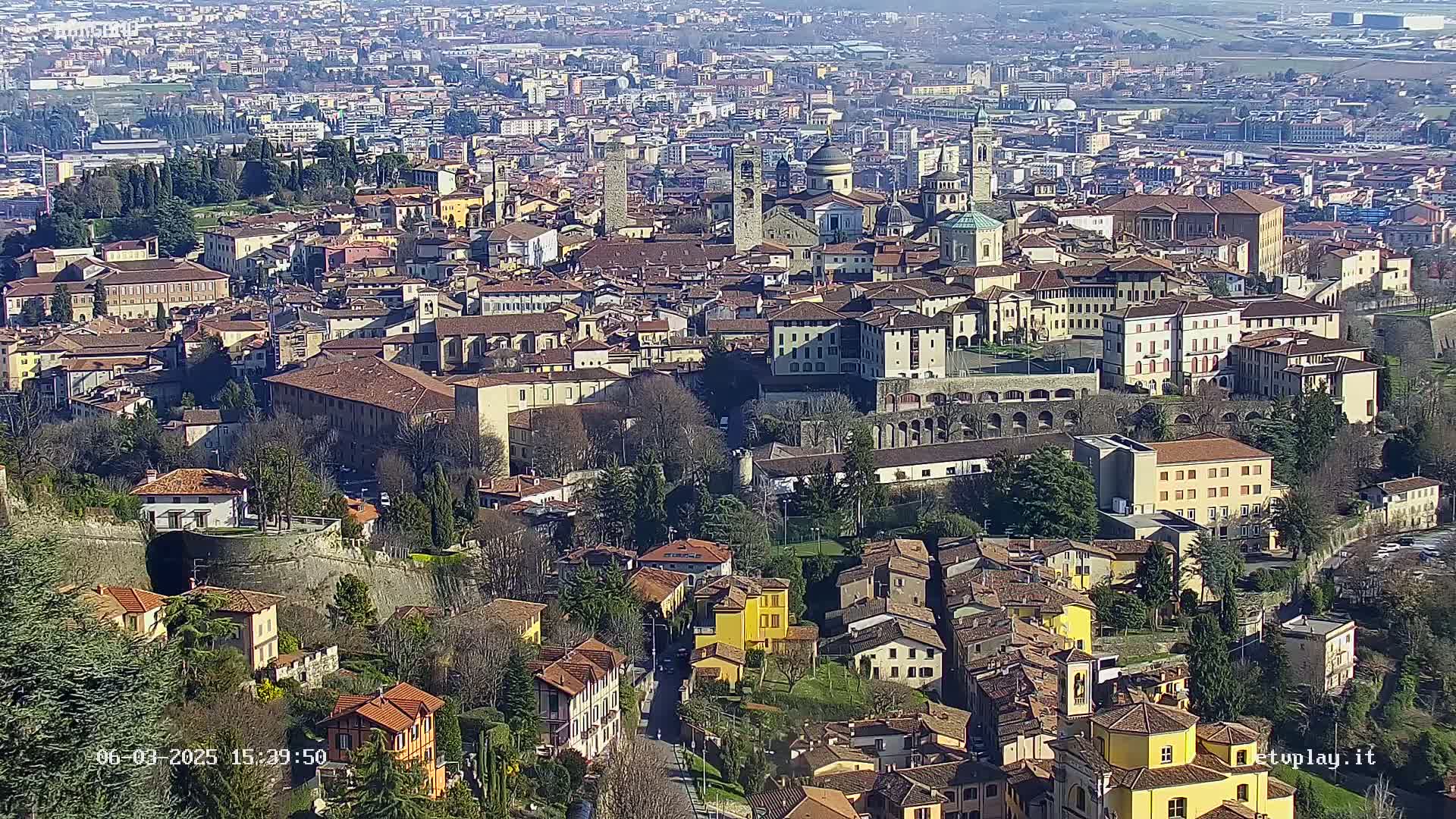 A sprawling city of red-roofed buildings and stone structures, nestled in a valley on a sunny day.