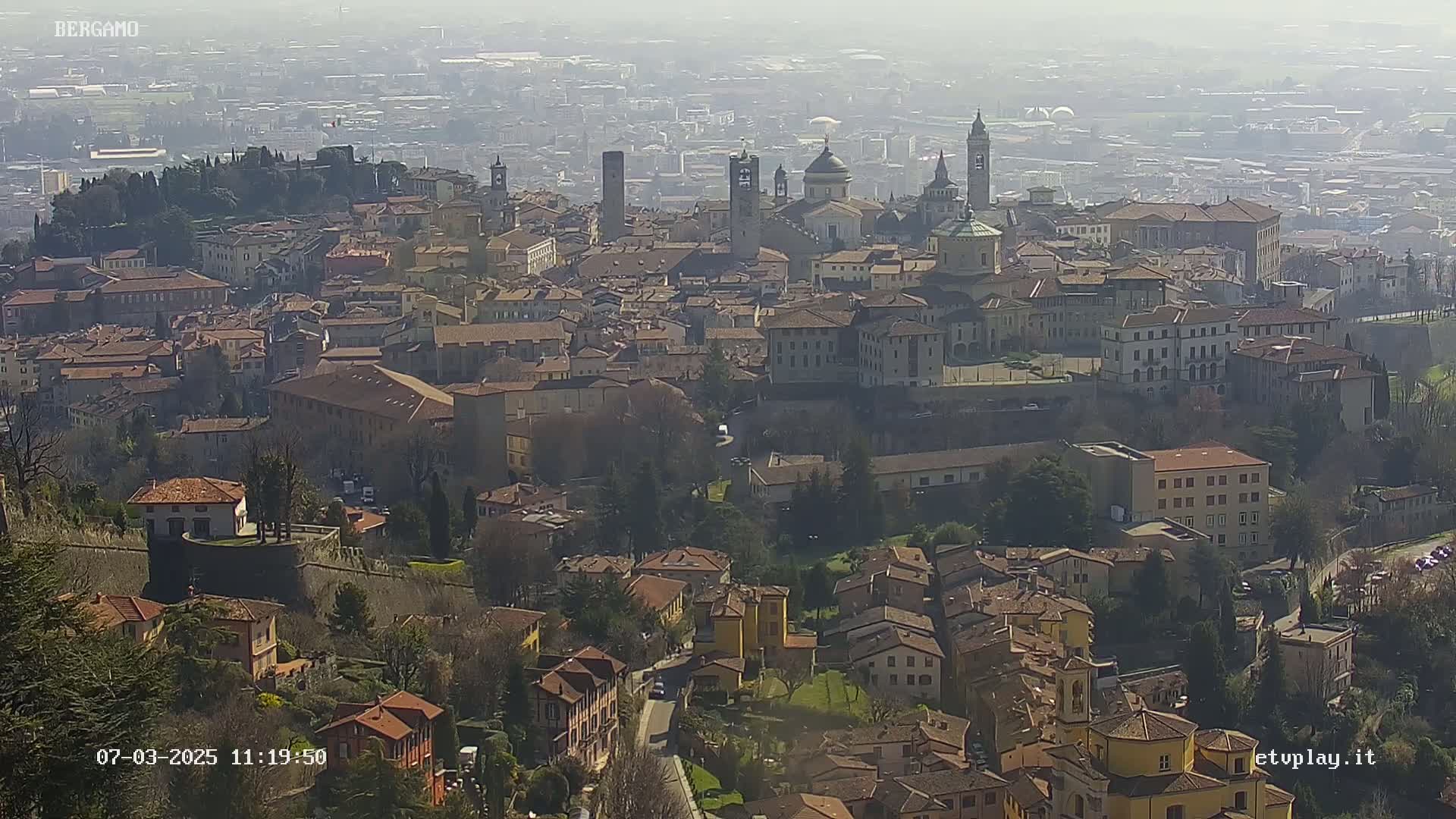 A hazy, sunny day reveals a sprawling cityscape of low-rise buildings, punctuated by several prominent churches and towers, nestled amongst green hills.