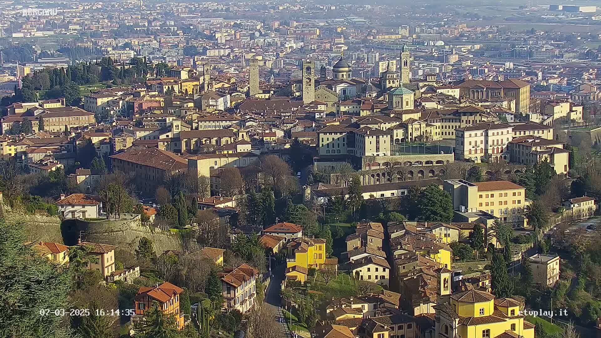 A sprawling cityscape of closely packed buildings, many with reddish-brown roofs, is viewed from a high vantage point on a sunny day.