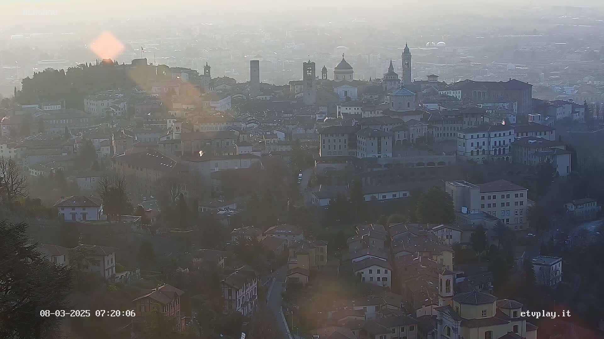A hazy sunrise illuminates a sprawling city with numerous buildings and several prominent church steeples.