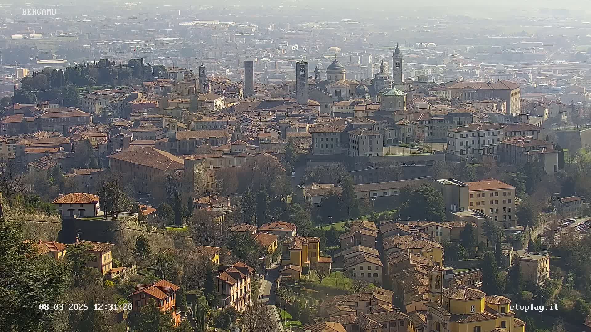 A hazy, sunny day reveals a sprawling cityscape of terracotta-roofed buildings nestled on a hillside, with taller structures and church domes visible in the distance.