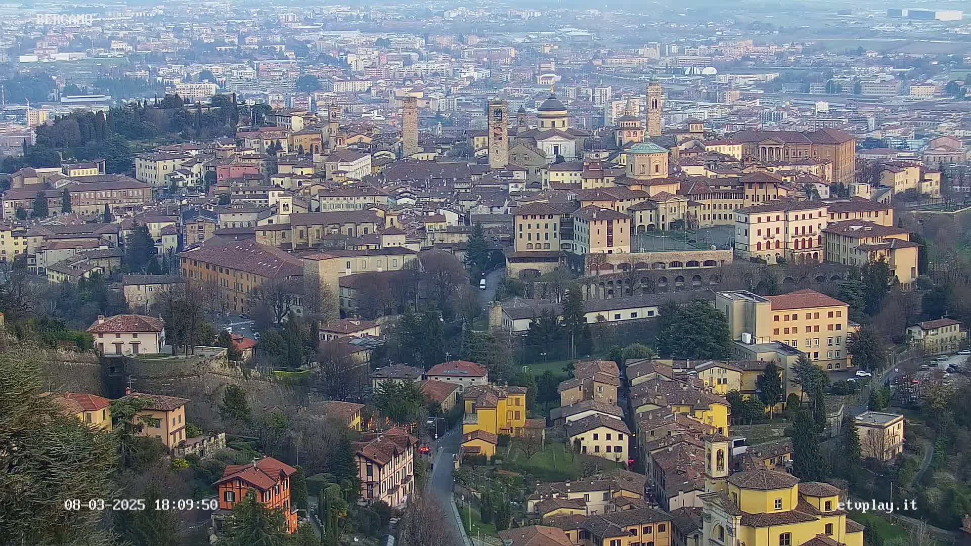 An aerial view shows a densely packed, historic city under a hazy sky, with buildings ranging from tall multi-story structures to smaller, closely situated houses.