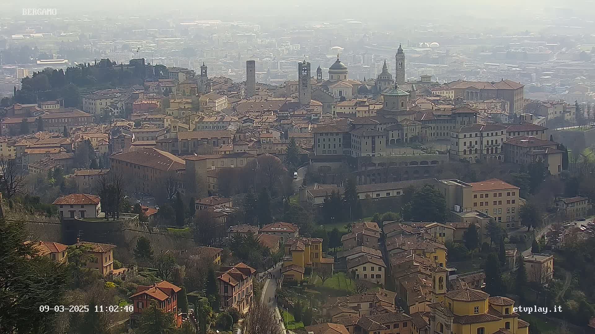 A hazy view of a sprawling city with numerous buildings, churches, and a hilltop fortress, under a foggy sky.
