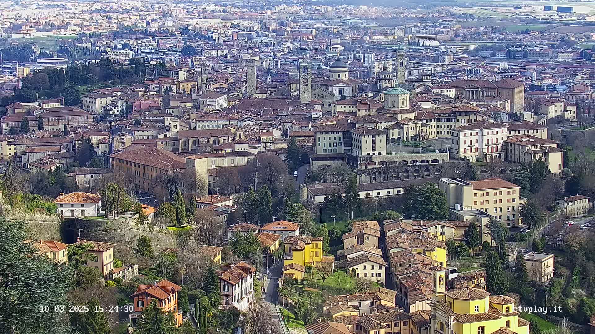 A sprawling city of closely packed, low-rise buildings with red-tiled roofs, partially surrounded by a stone wall, on a partly sunny day.