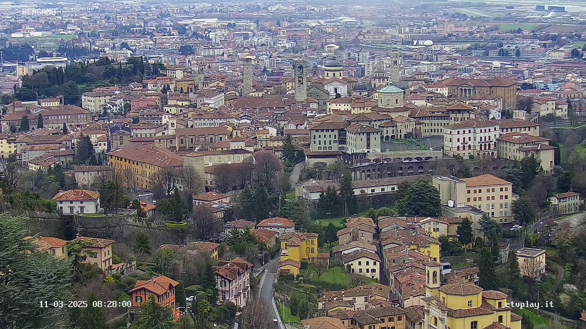 A sprawling city of closely packed, terracotta-roofed buildings stretches across a hillside under an overcast sky.