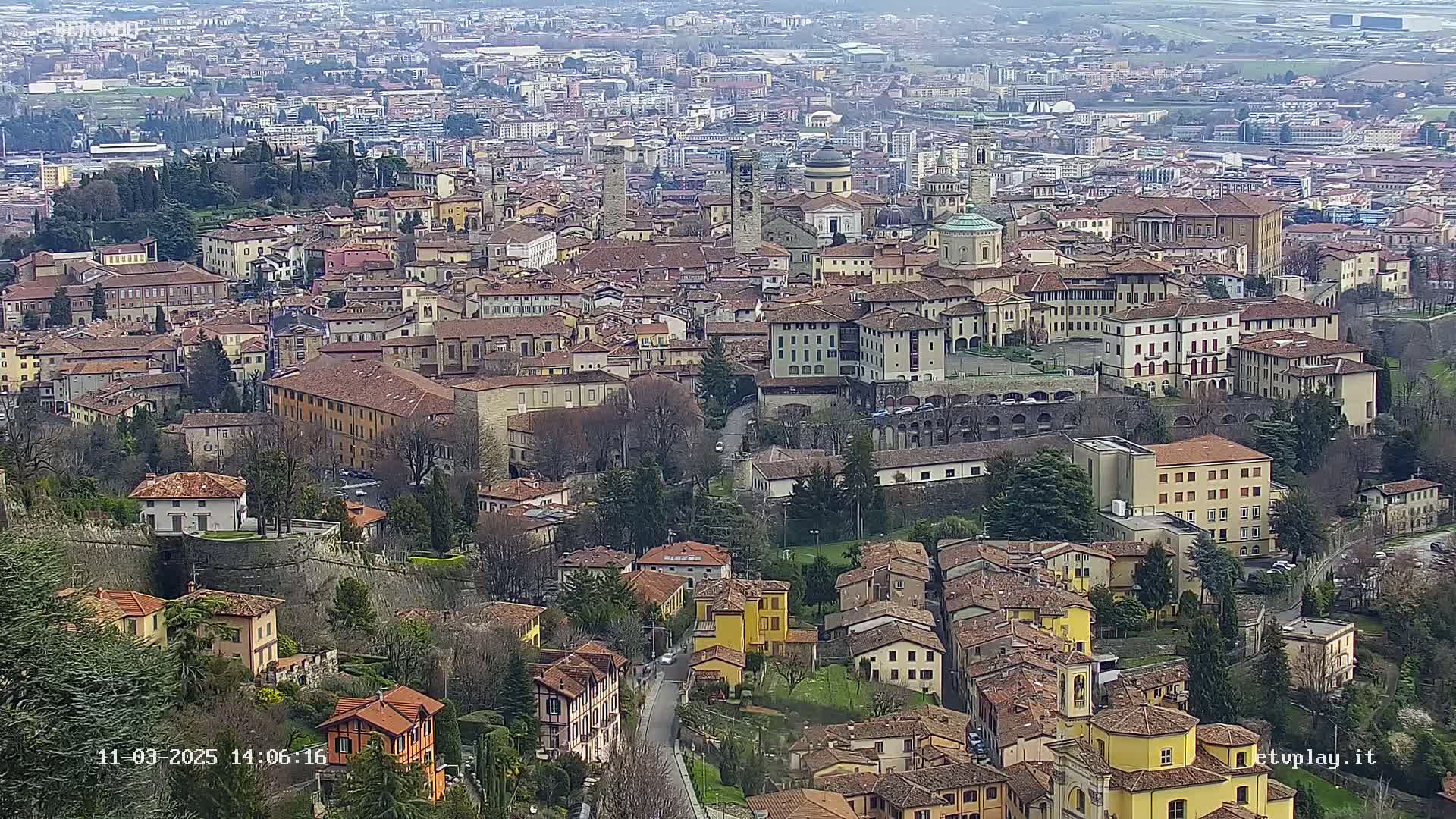 A sprawling city of closely packed, low-rise buildings with red-tiled roofs sits under a cloudy sky.