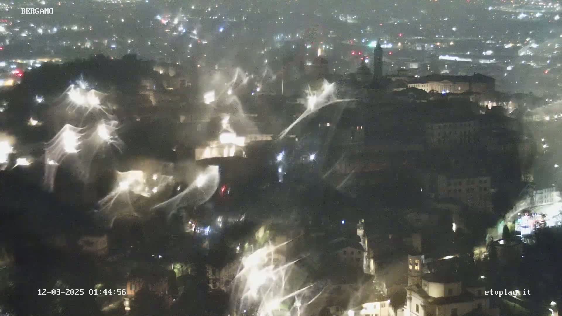 A nighttime, high-angle view shows a city illuminated with many lights, overlaid with streaks of what appears to be heavy snow or intense rain.