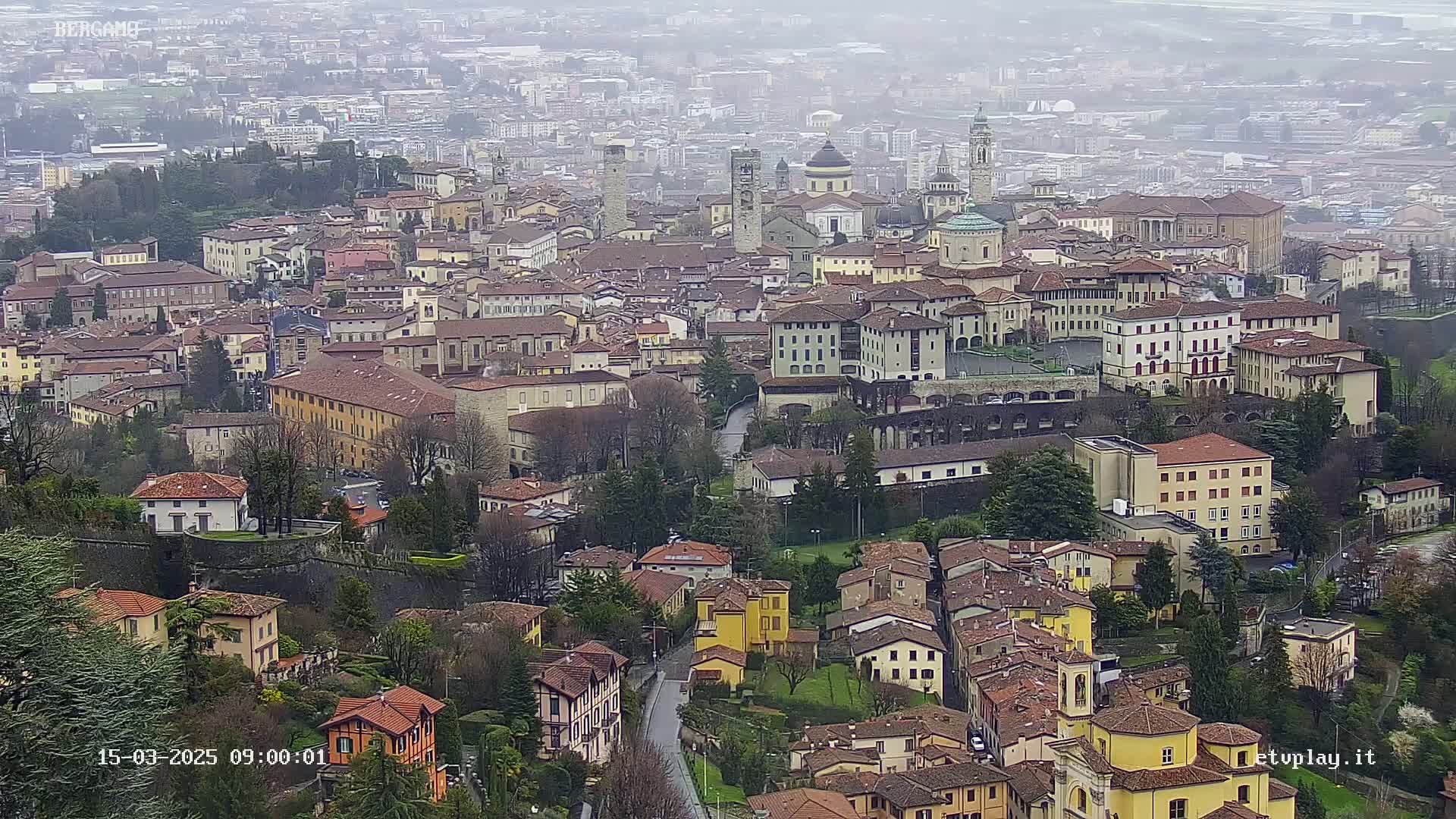 A sprawling city with numerous buildings of varying heights and styles is viewed from above on an overcast day.