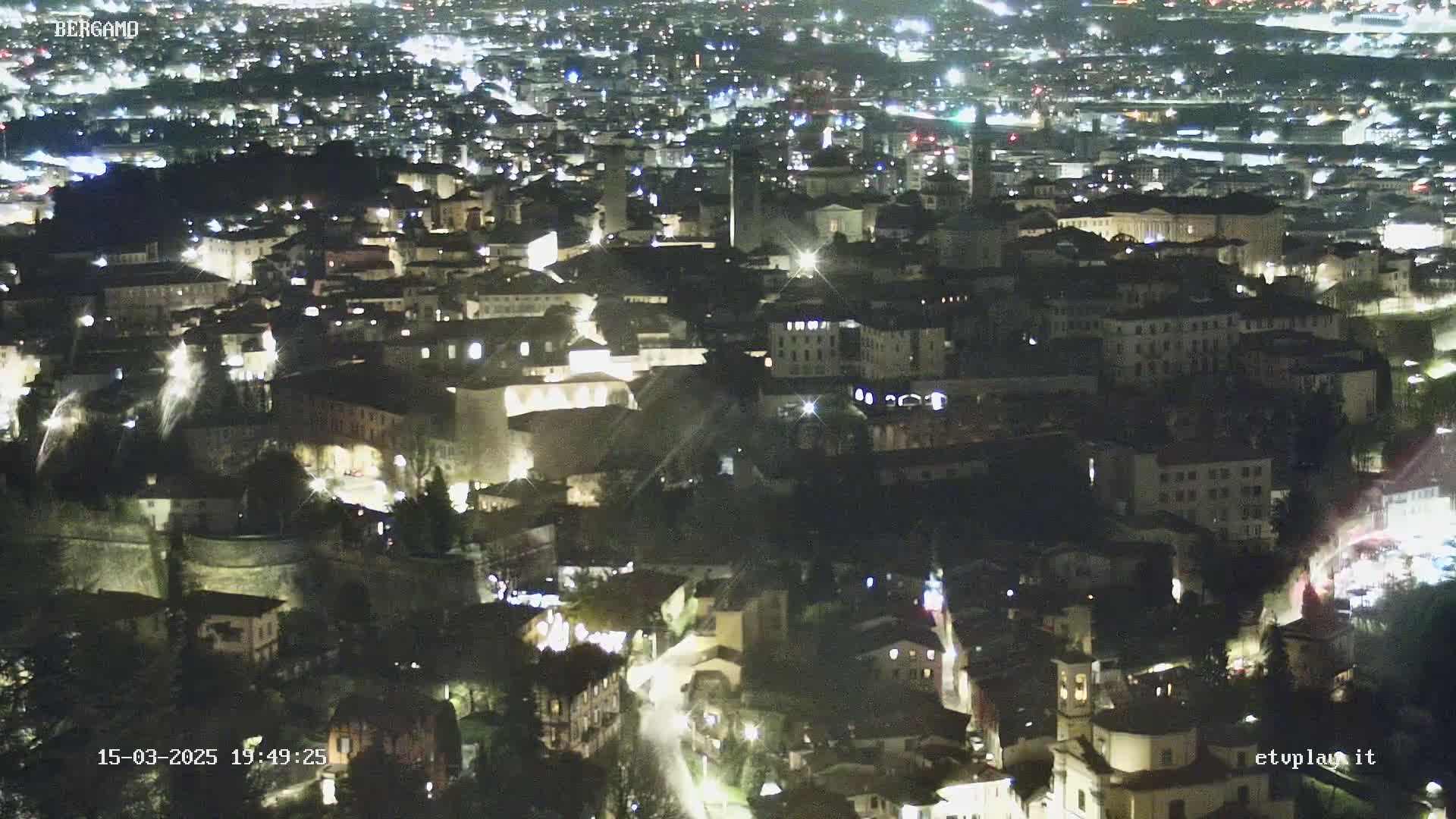 A nighttime aerial view of a city illuminated by streetlights, showing a dense collection of buildings nestled within a hilly landscape.