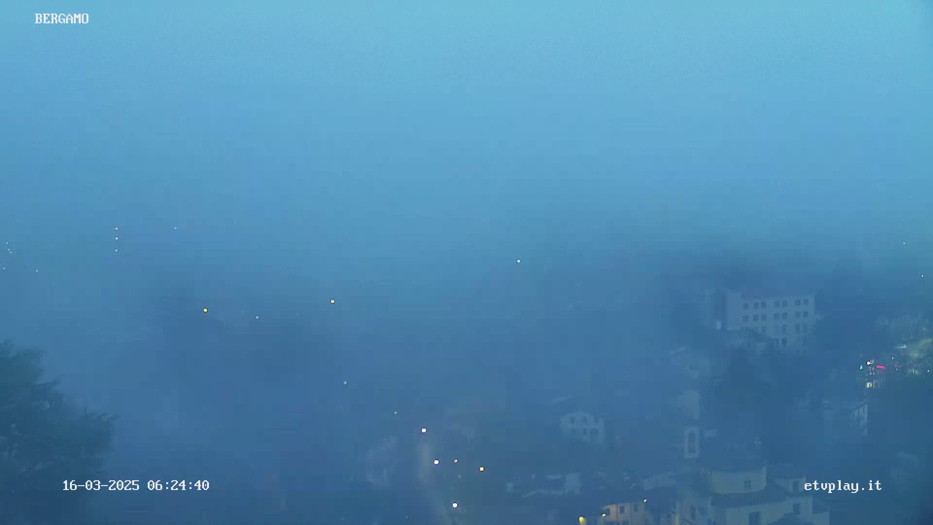 A foggy, twilight view shows a town partially obscured by thick fog, with a few lights visible.