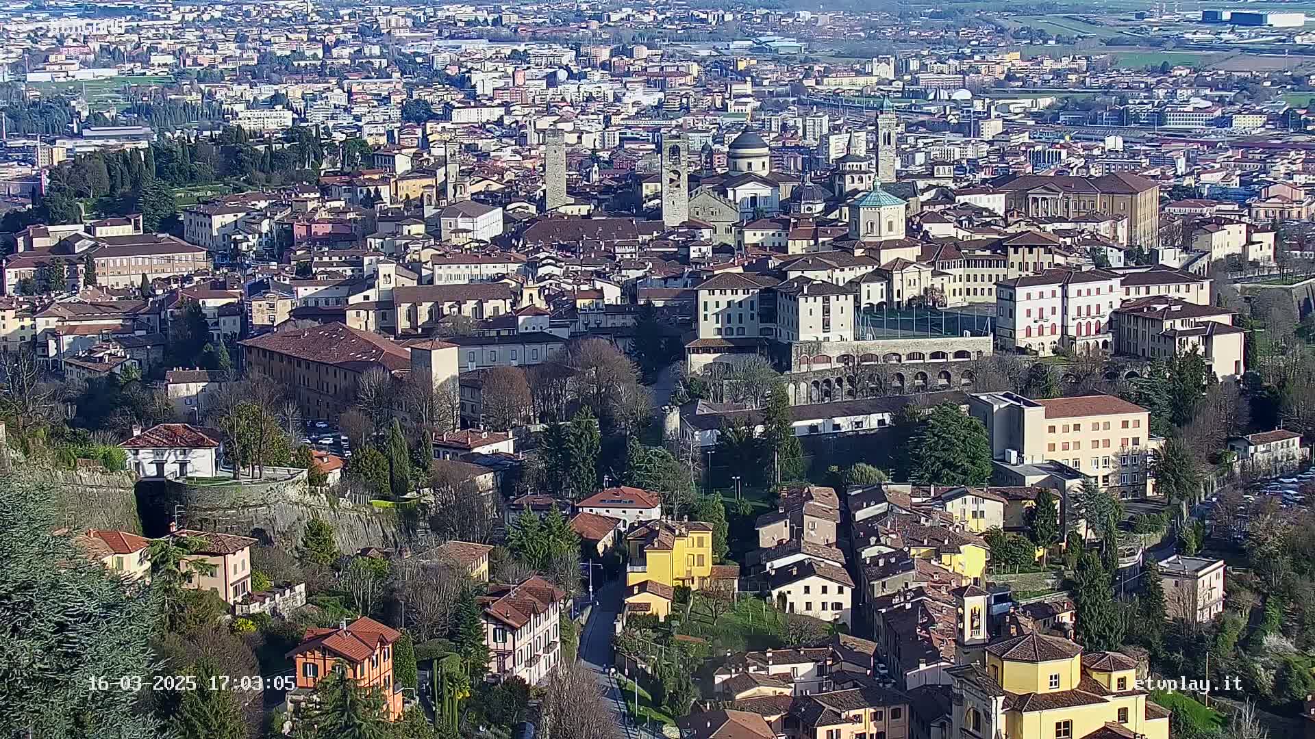 A sprawling town of closely clustered buildings, many with reddish-brown roofs, is visible from a high vantage point on a sunny day.