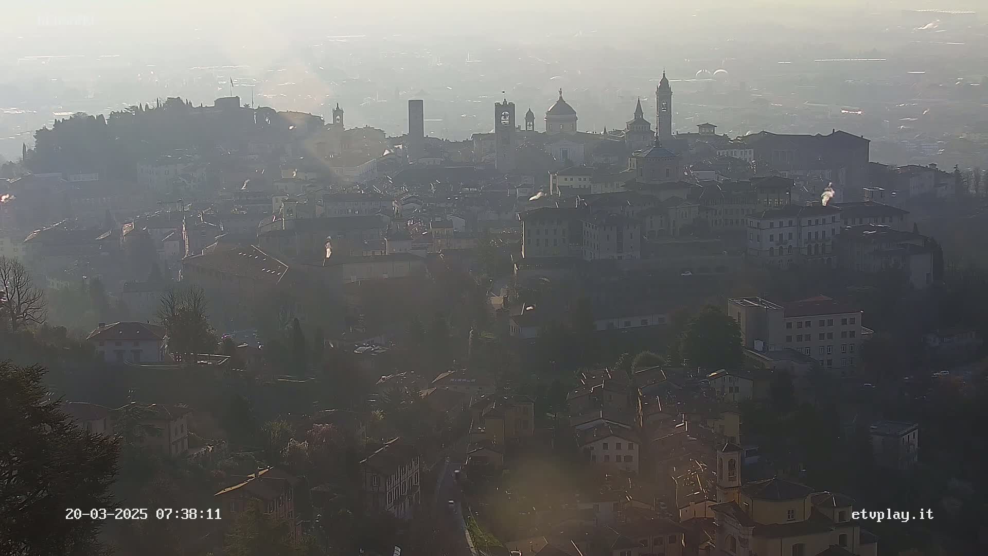 A hazy sunrise illuminates a sprawling town nestled in a valley, with numerous buildings and church steeples visible in the distance.
