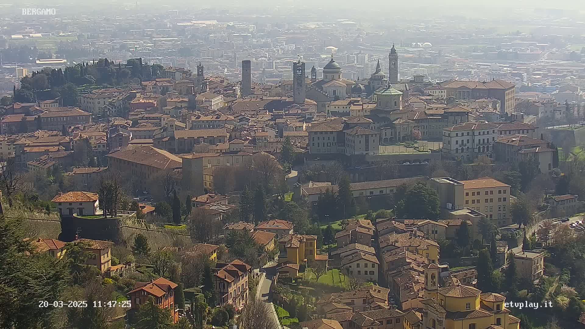 A hazy, sunny day reveals a sprawling city of terracotta-roofed buildings, punctuated by churches and towers, nestled in a hilly landscape.